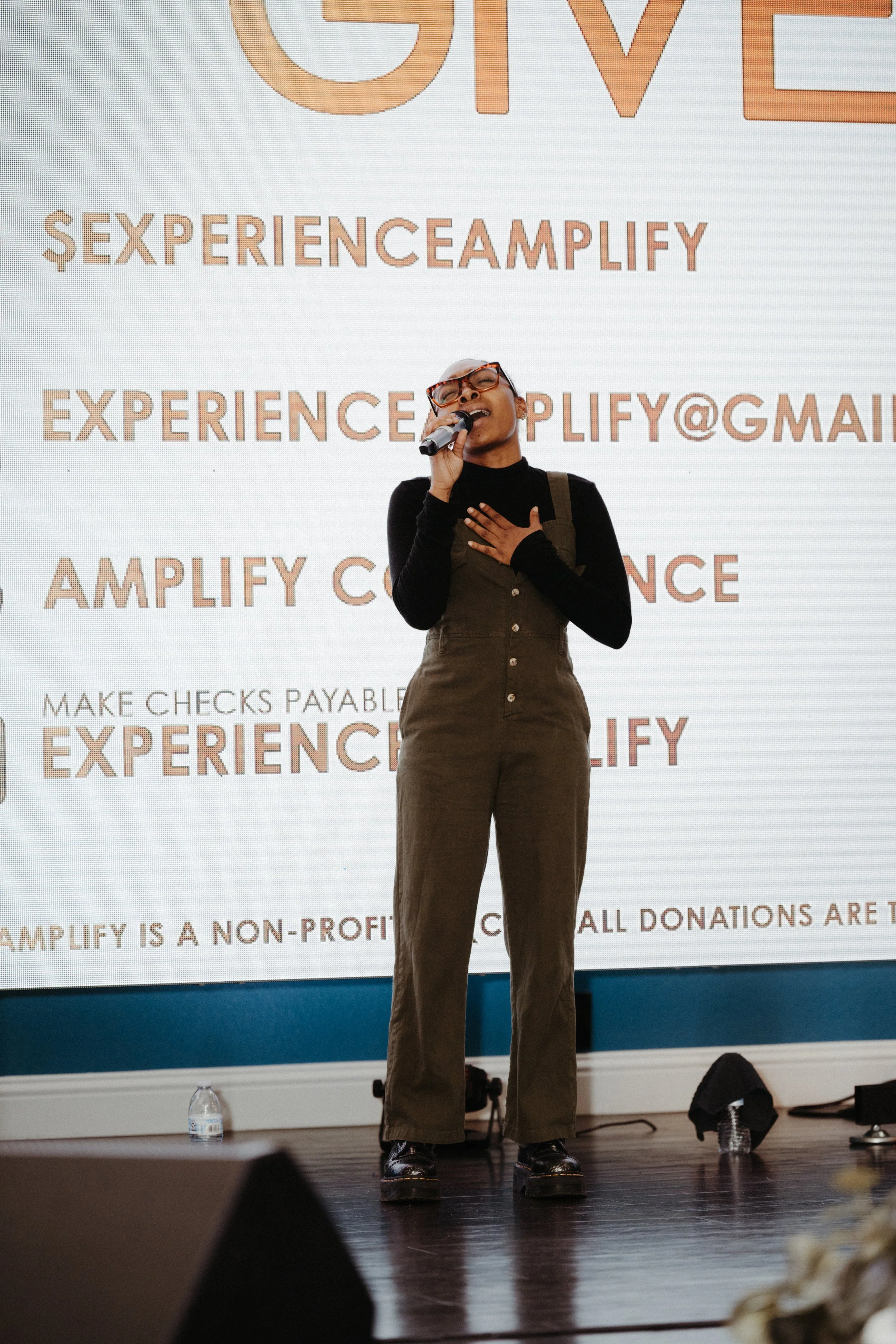A woman singing into a microphone on stage, wearing glasses, a black shirt, and olive-green overalls, with a large presentation screen behind her.