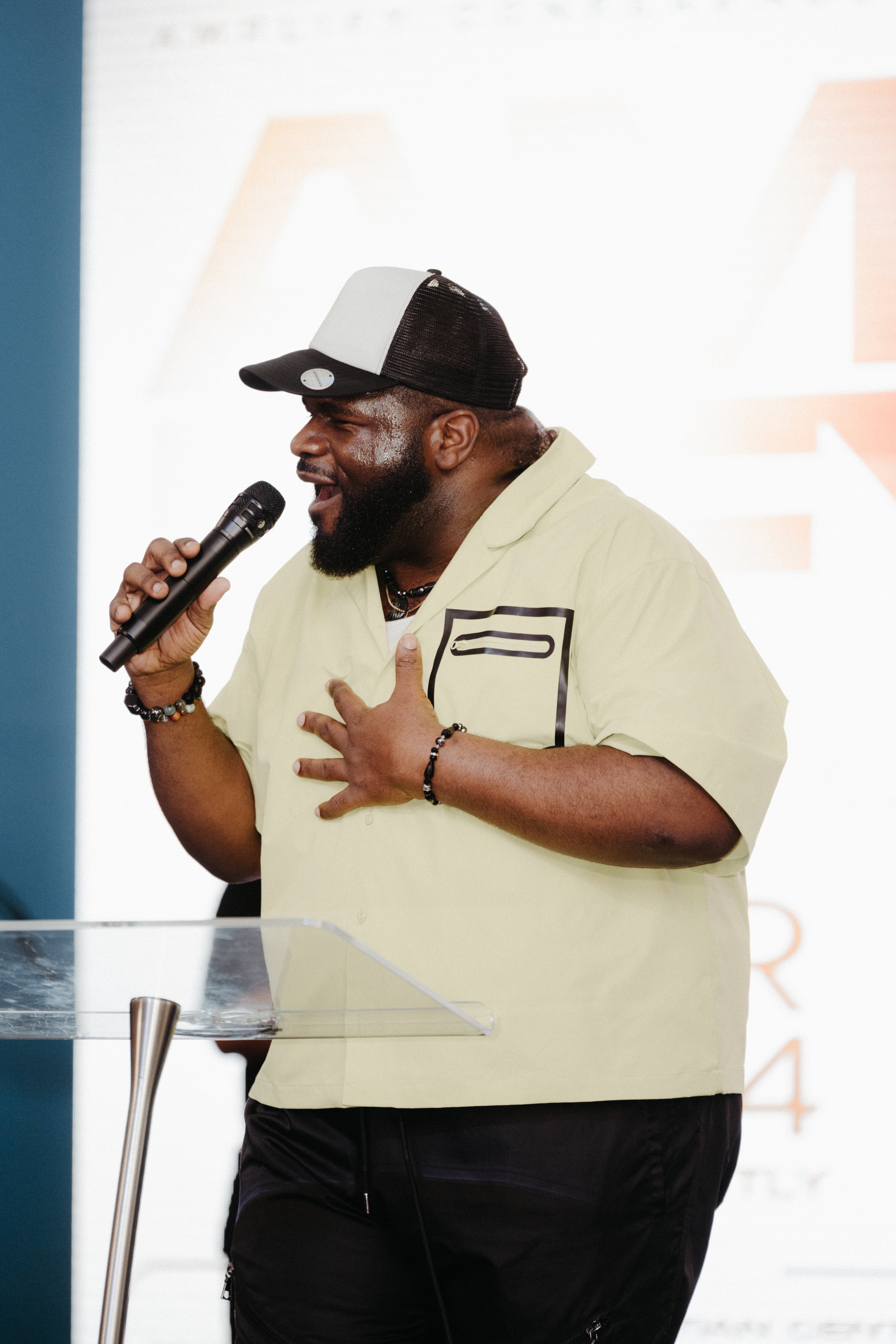 A man with a beard wearing a cream-colored shirt and black pants, holding a microphone, singing or speaking passionately, with his left hand on his chest, standing behind a clear lectern, in front of a white screen with some partially visible large text.