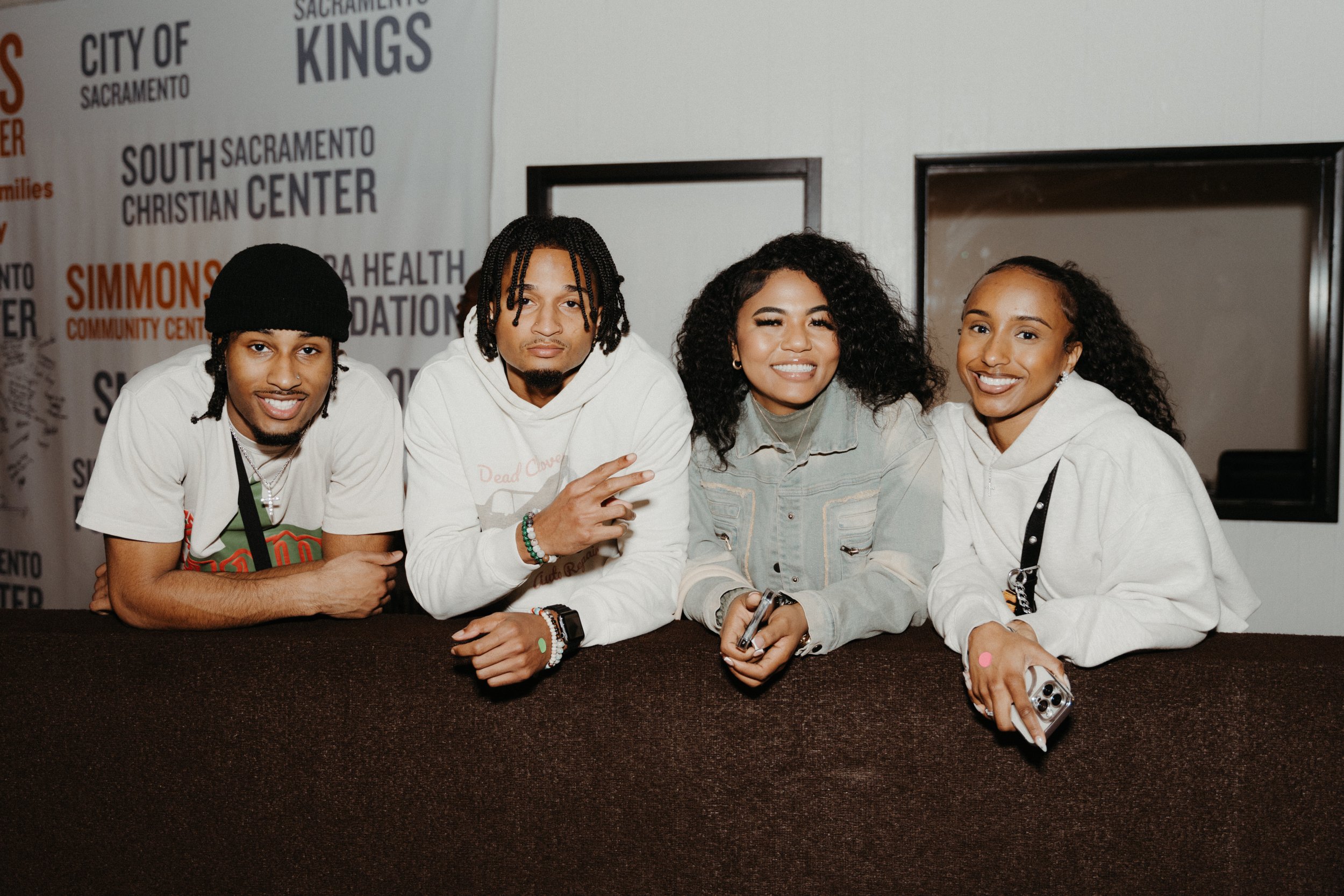 Four young adults sitting behind a table, smiling, with a wall featuring various texts and logos in the background.