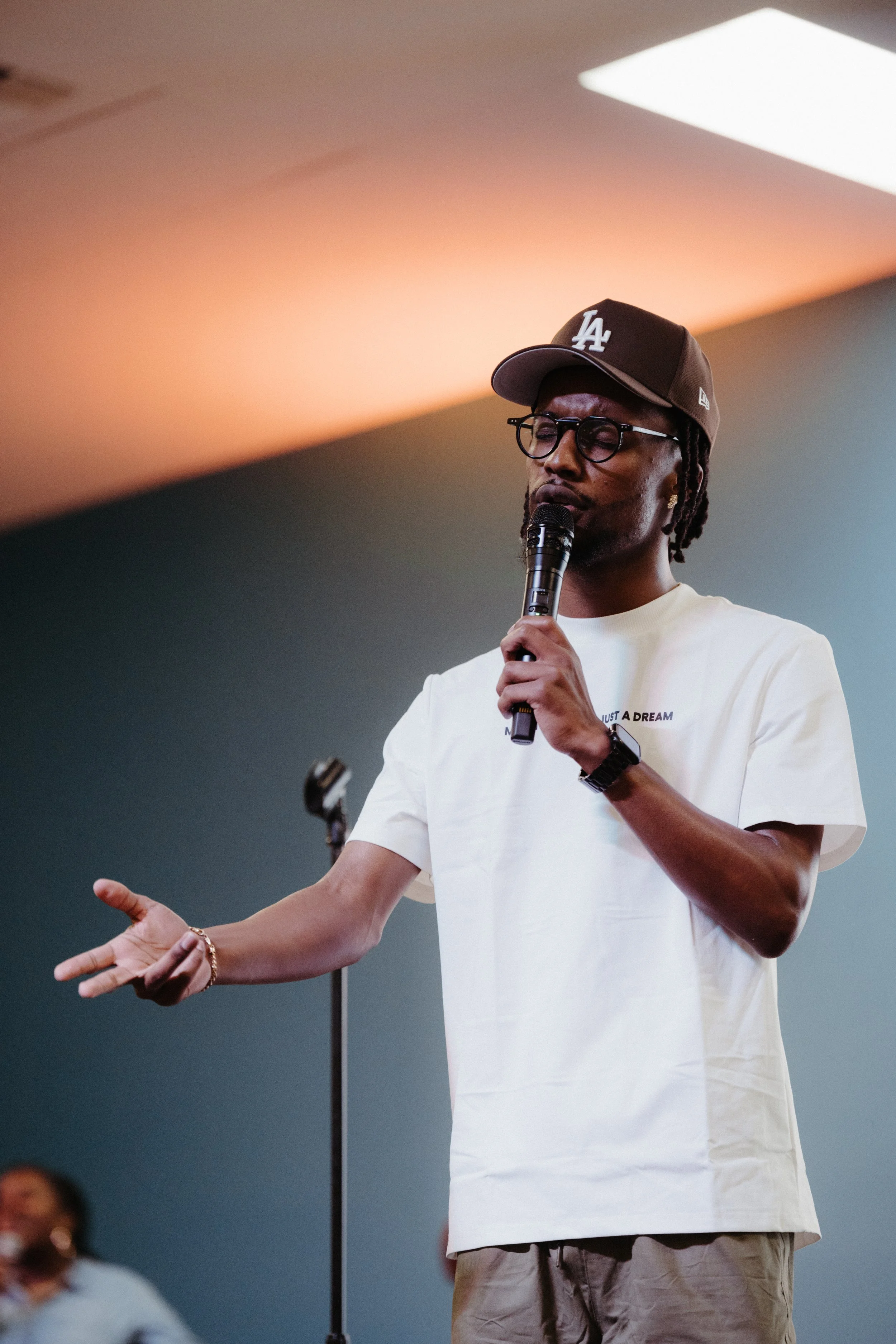 A man wearing glasses and a baseball cap speaking into a microphone during a presentation or speech, gesturing with his left hand.