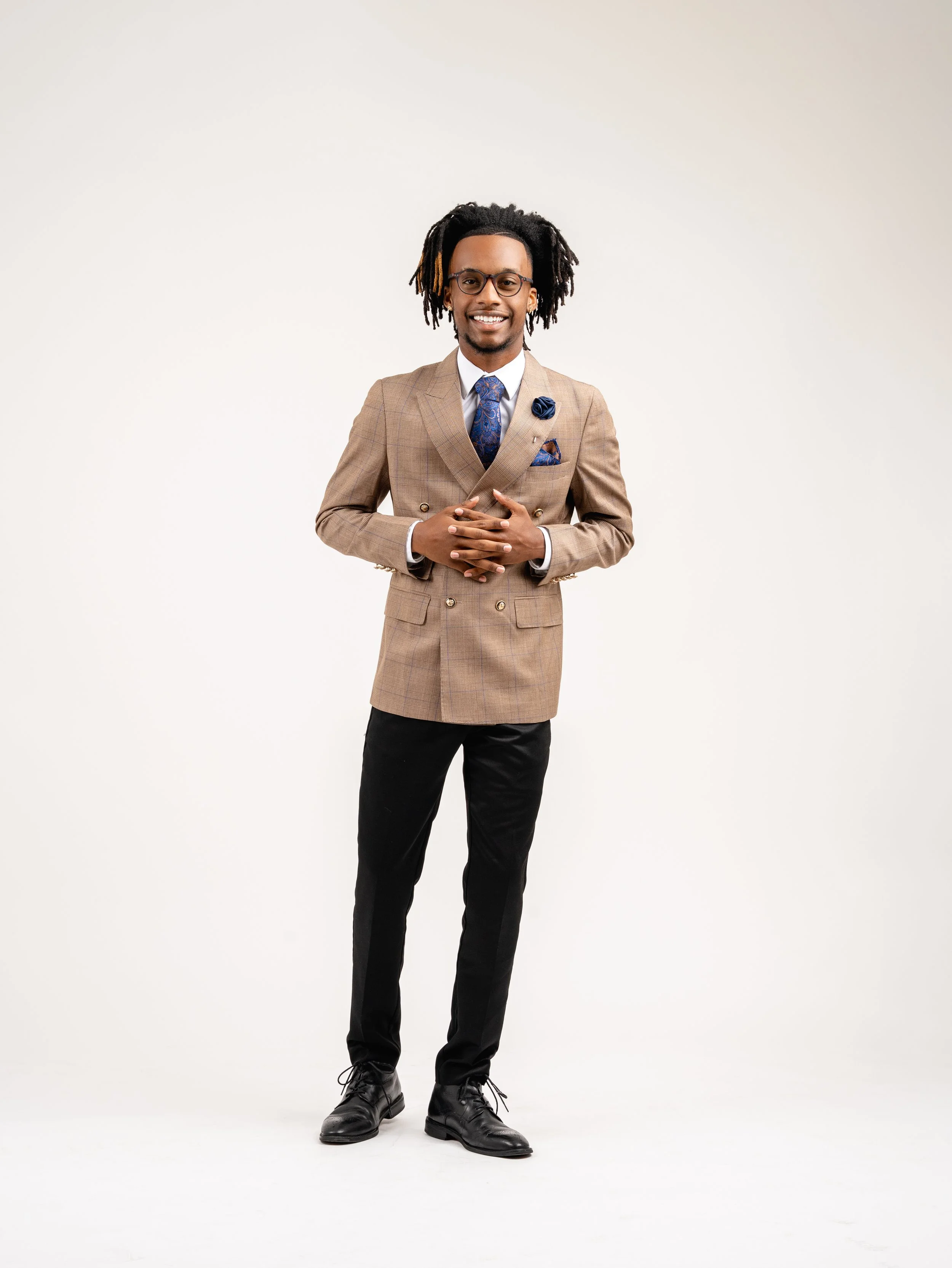 A man in a beige double-breasted blazer, white shirt, and tie, standing against a plain white background, smiling with hands clasped in front.