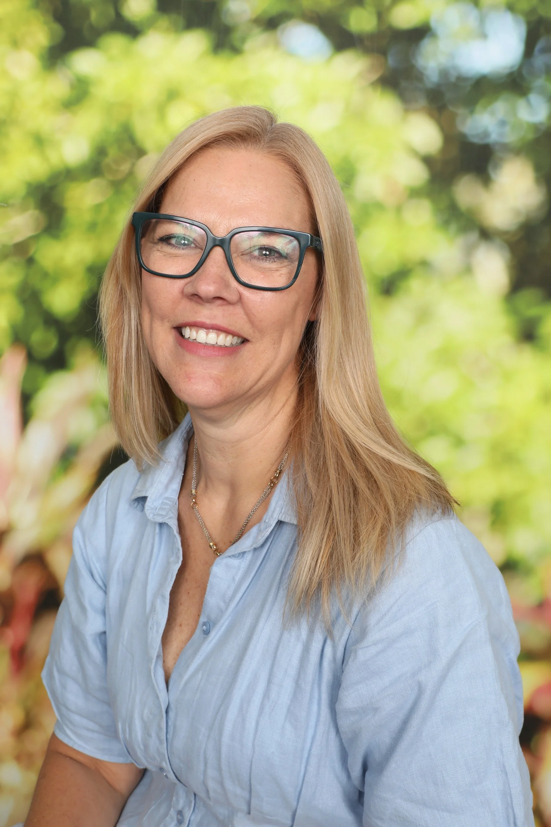 A woman with blonde hair, glasses, and a blue shirt smiling outdoors with a blurred green and brown background.