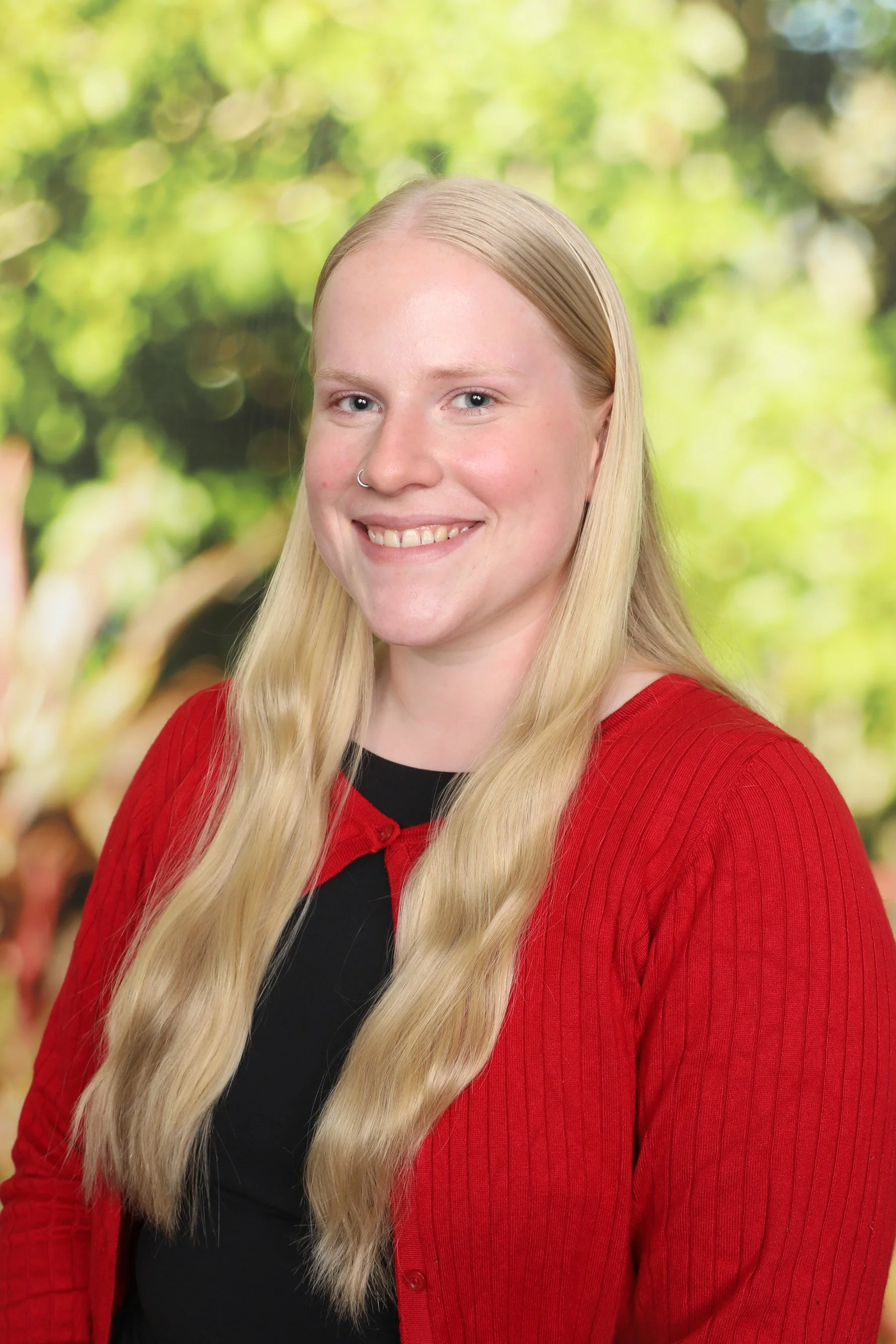 Portrait of a young woman with long blonde hair, wearing a red cardigan, smiling, with a blurred green outdoor background.