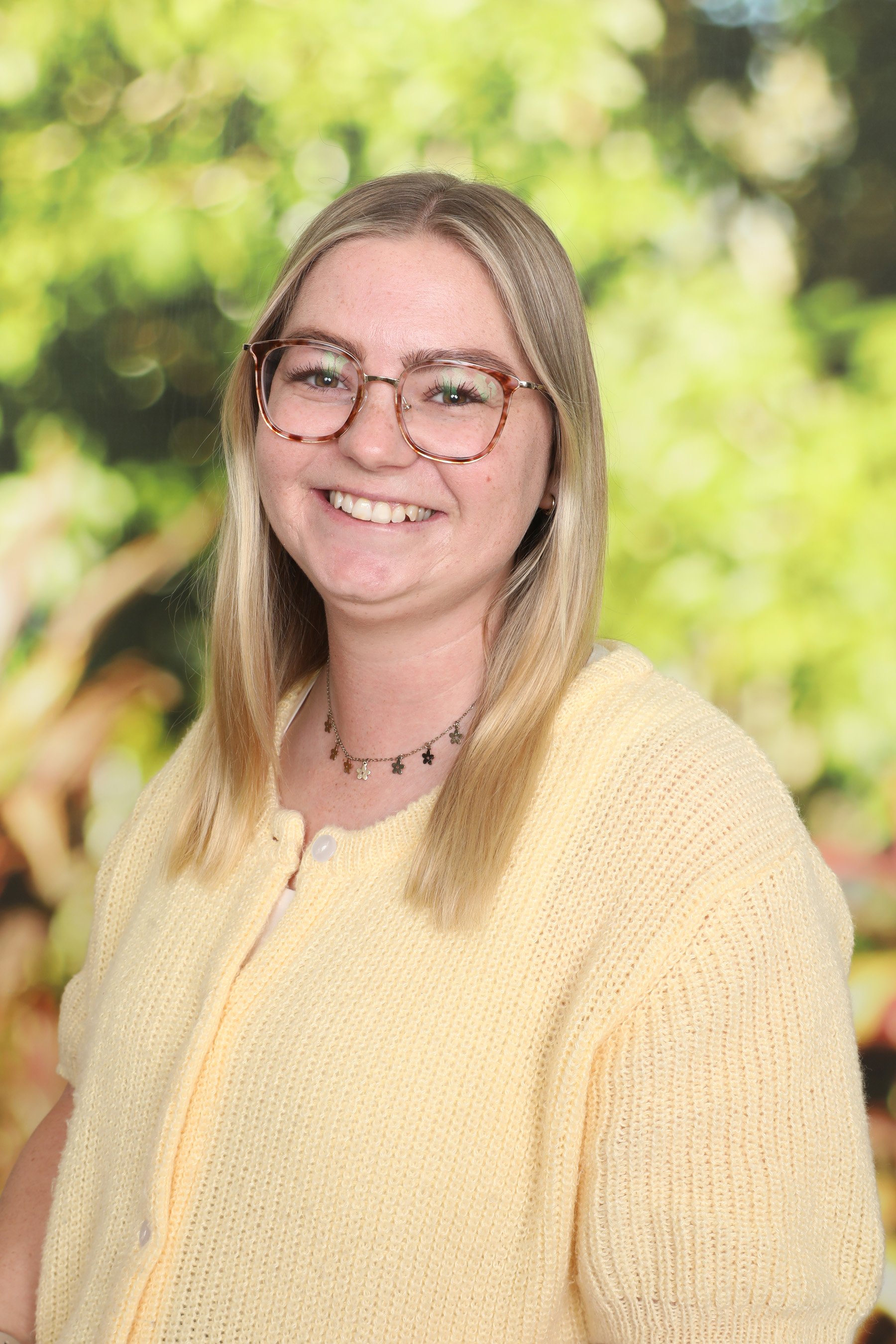 Photo of a young woman with blonde hair, glasses, and a cheerful smile, wearing a yellow sweater and a necklace with star-shaped charms, standing outdoors with a blurred green and yellow background.