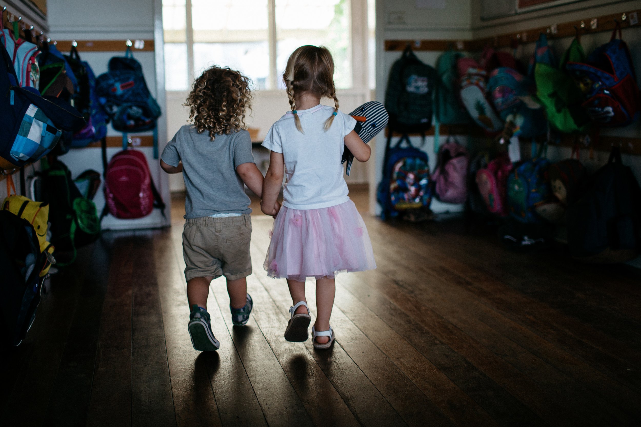 Two young children, a boy and a girl, walking down the hallway of a daycare with backpacks hanging on hooks on both sides of the hallway.