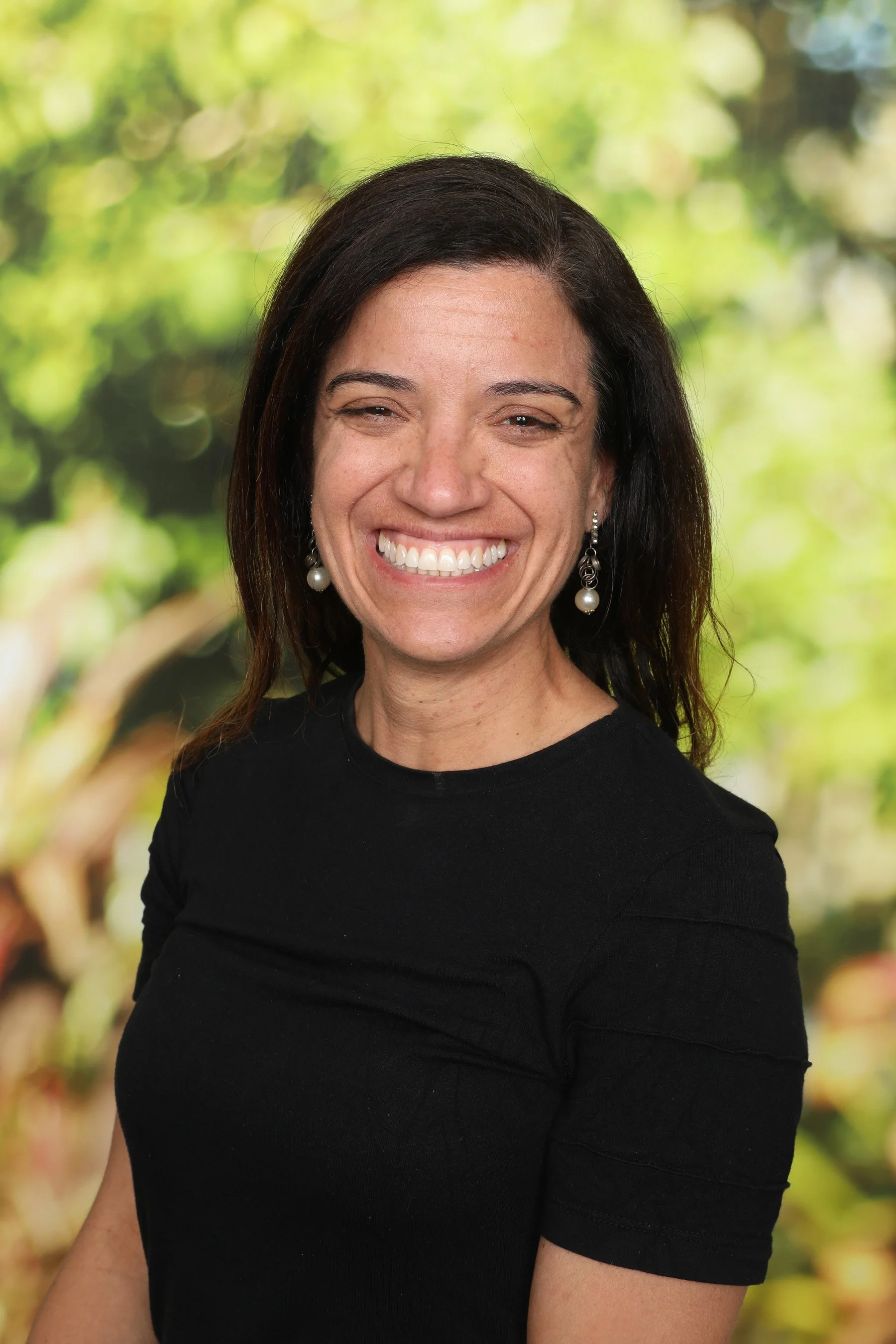 A woman with dark brown hair, wearing a black shirt and pearl earrings, smiling outdoors with a blurred green foliage background.