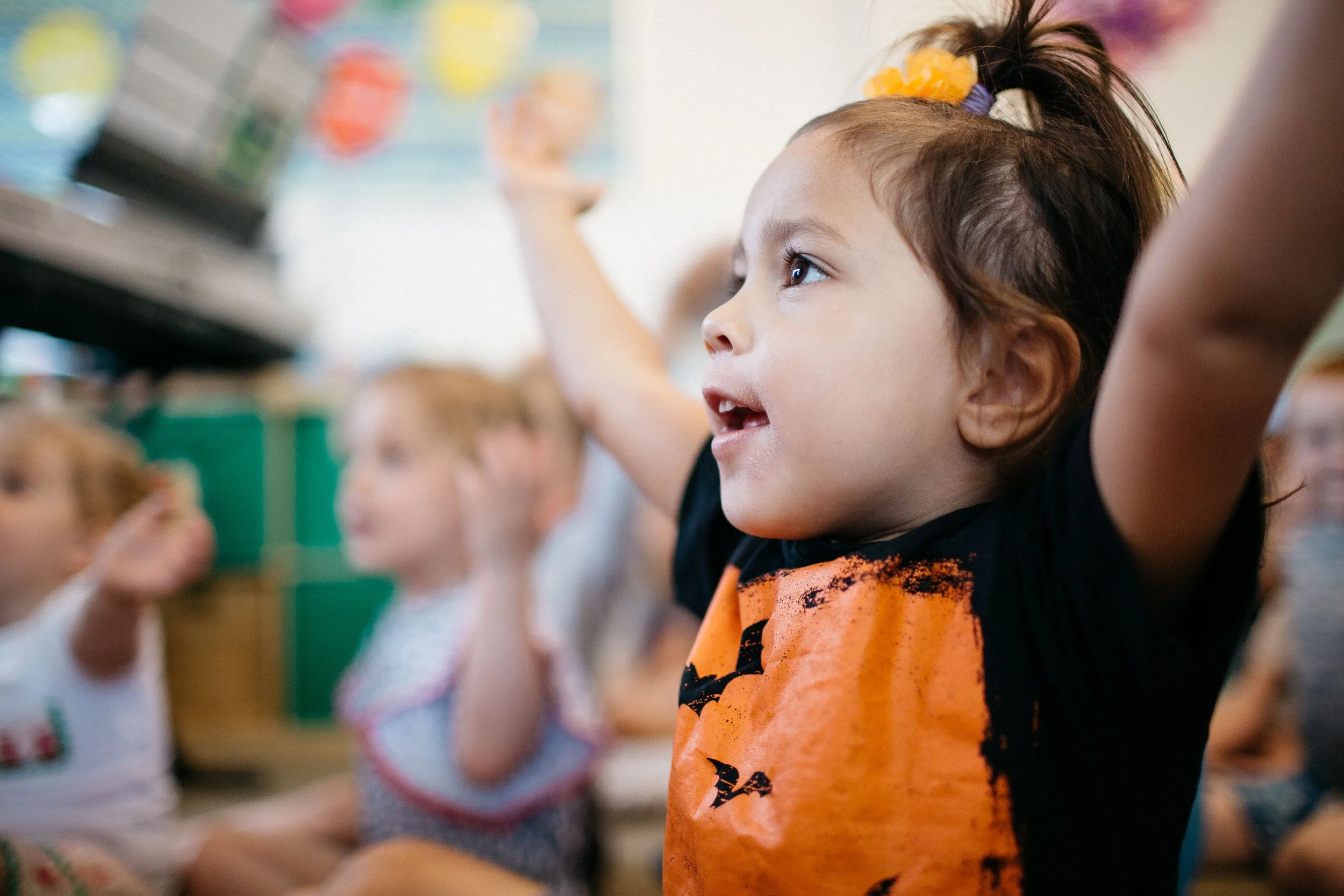 A young girl in a classroom, raising her hand, wearing an orange shirt with a black design, with other children visible in the background.