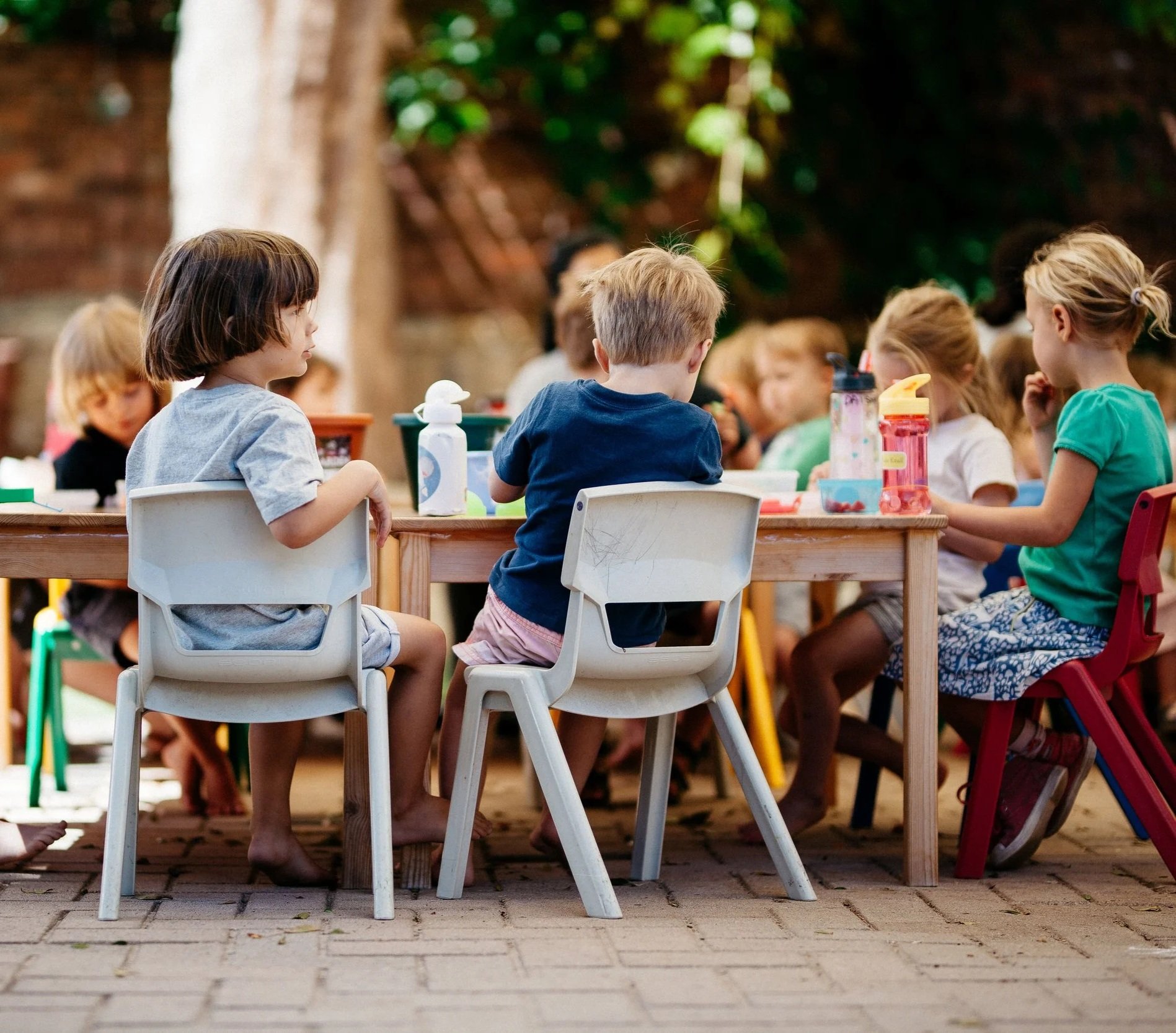 Children sitting at a long wooden table outdoors, engaged in eating or activities, with water bottles and cups on the table, in a garden or backyard setting.