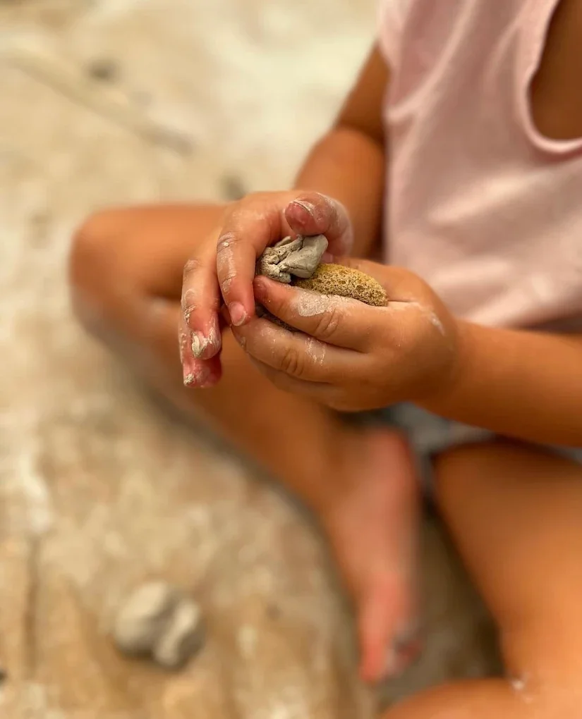 Child sitting on the floor playing with clay, hands covered in clay, holding a small clay sculpture.
