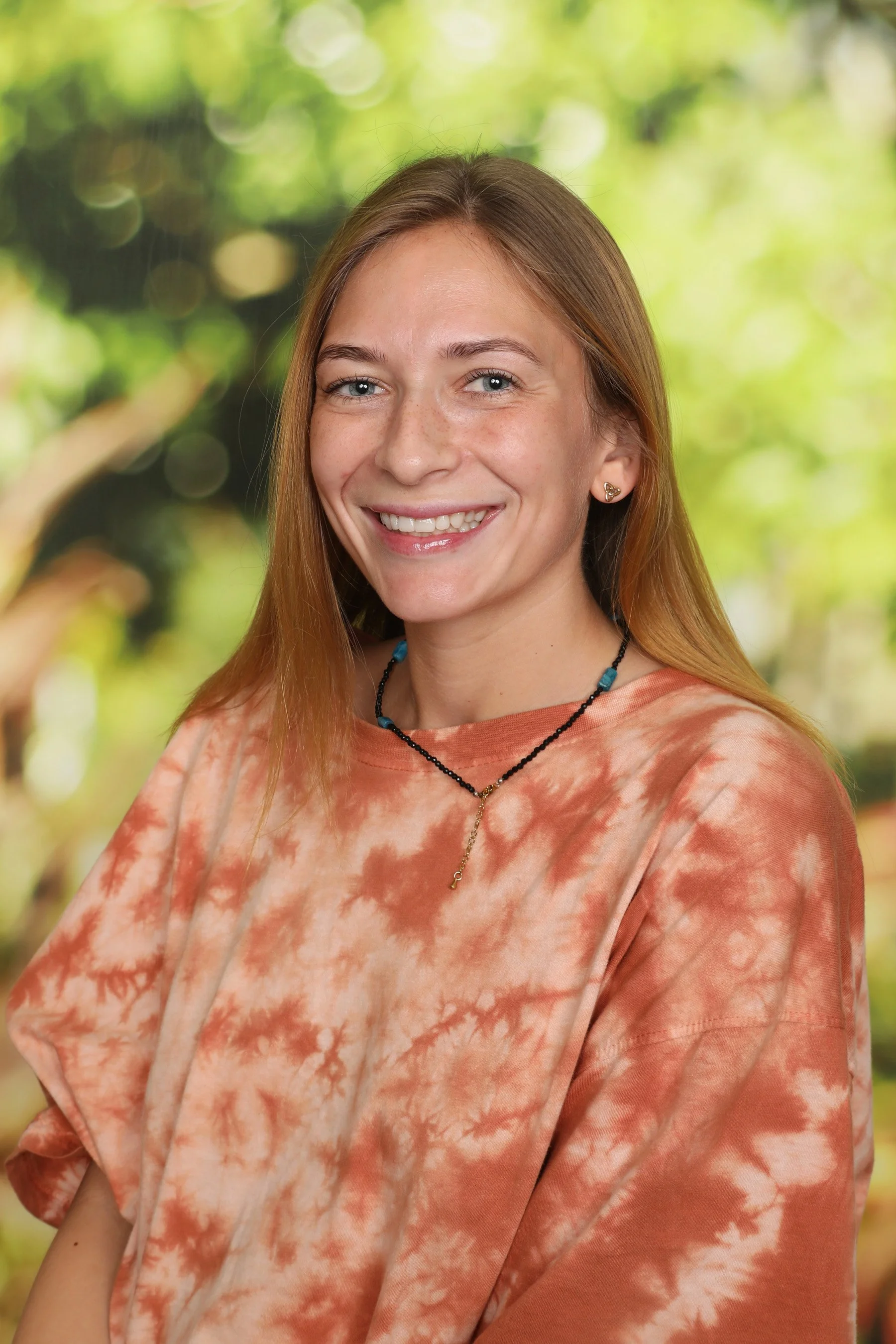 A young woman with long, straight, light brown hair and blue eyes, smiling while wearing a tie-dye orange and beige shirt and a beaded necklace, with a blurry green outdoor background.