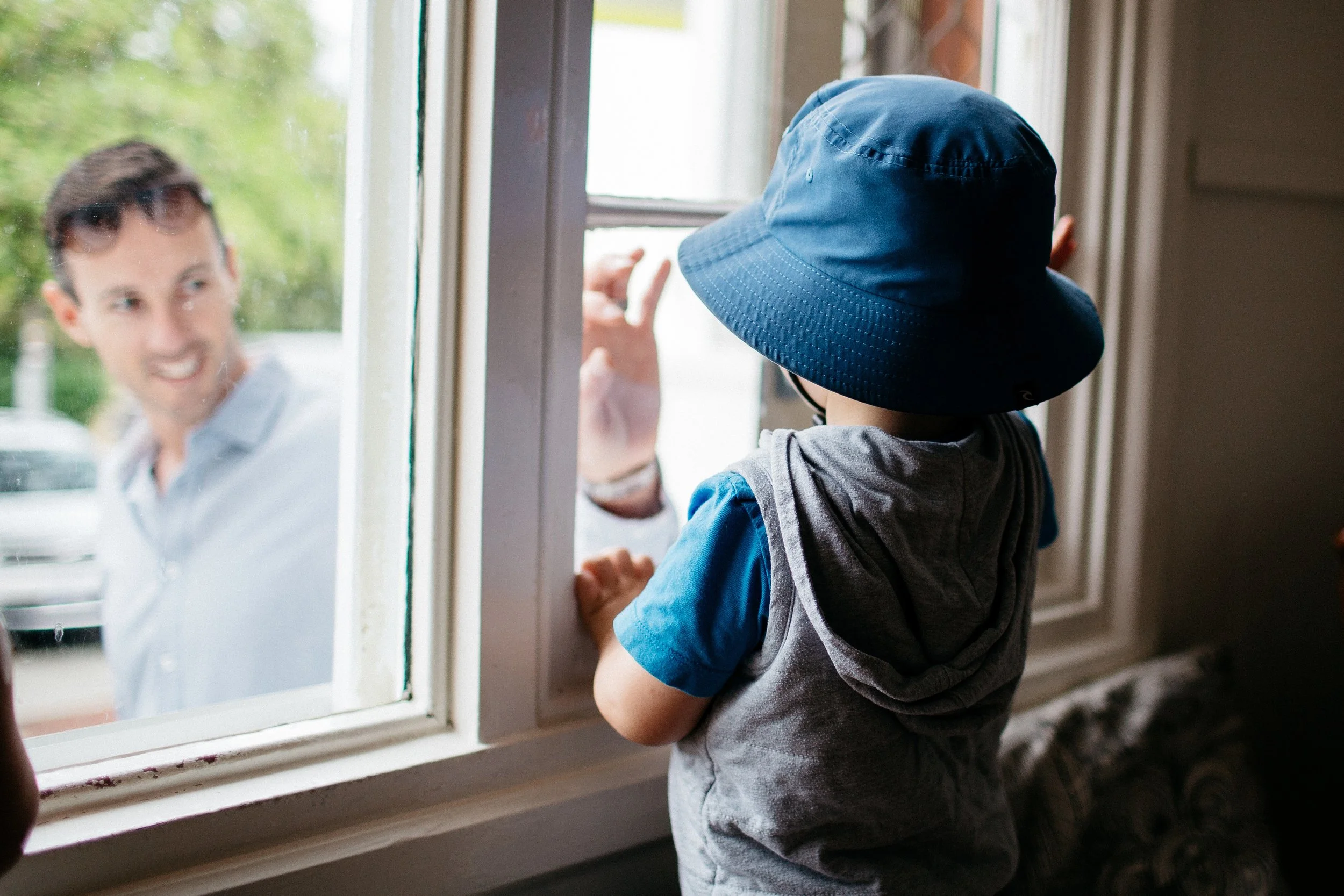 A young boy wearing a blue hat and a hoodie looks out a window at an adult man outside, who is smiling and wearing sunglasses.