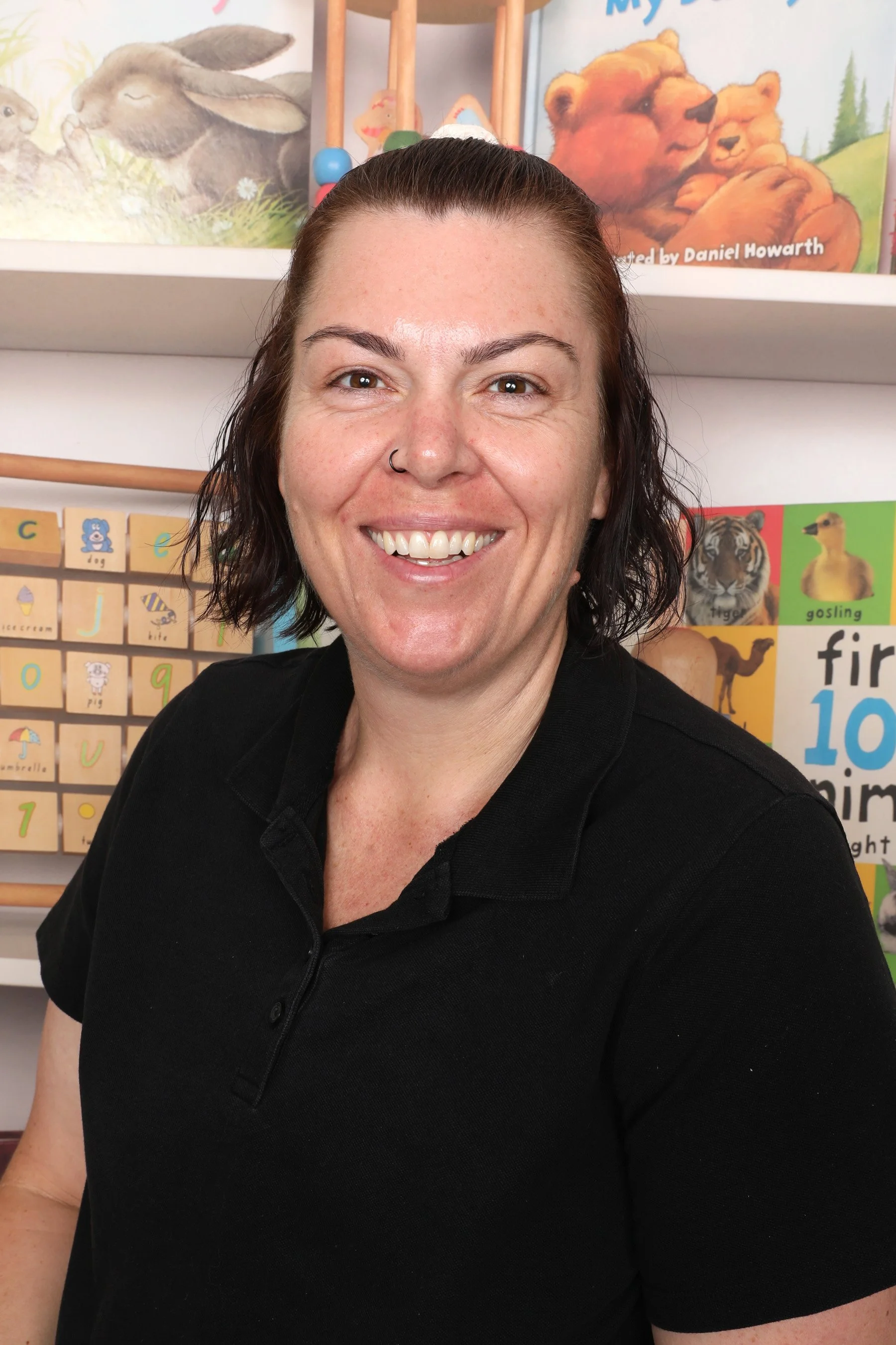 A woman smiling at the camera, wearing a black polo shirt, standing in a classroom with educational posters and picture cards of animals and alphabet letters on the wall behind her.