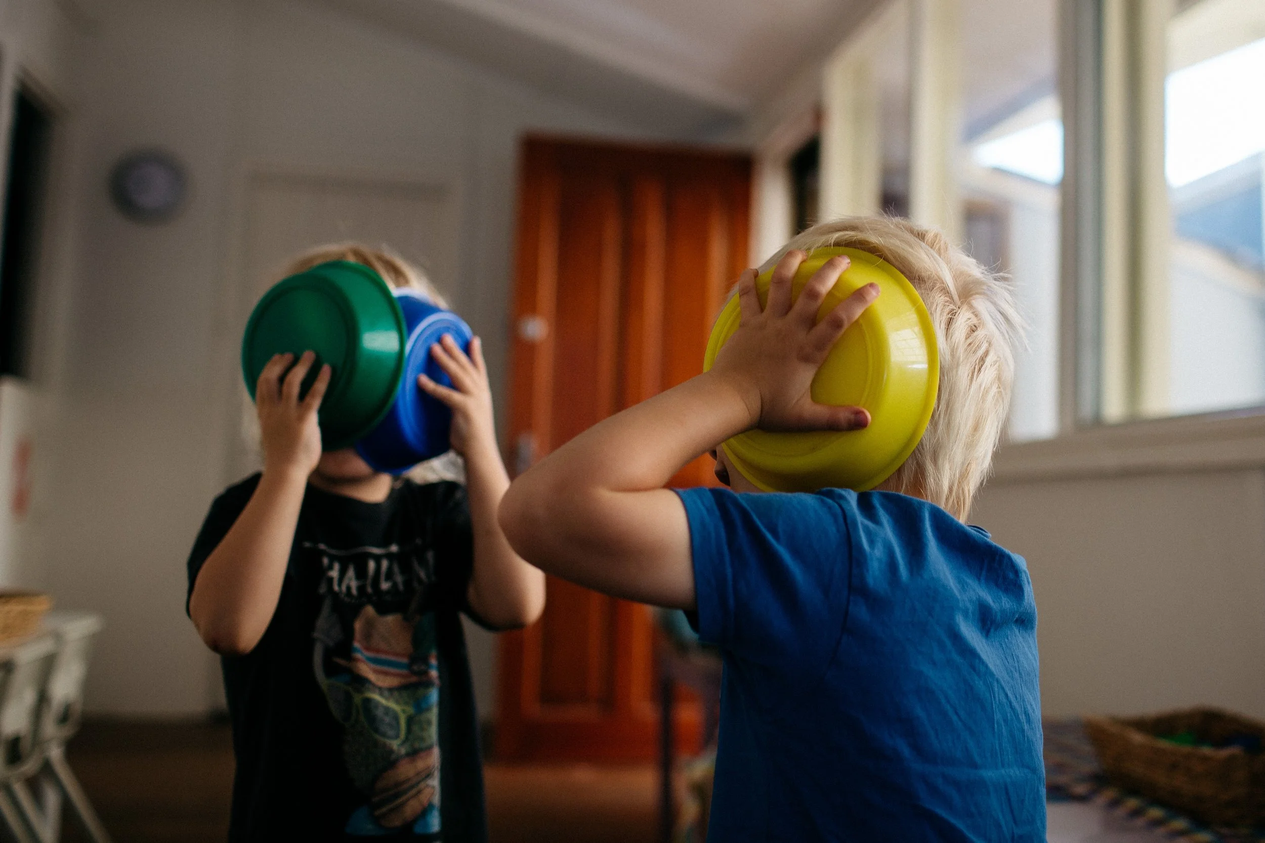 Two young children playing indoors, covering their ears with colorful plastic bowls.