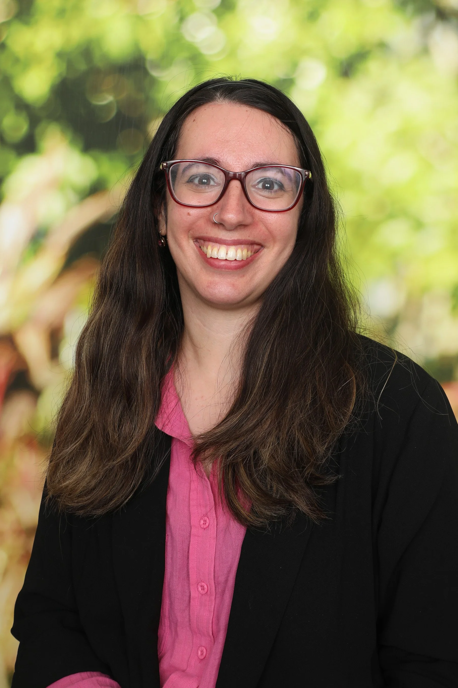 A woman with long brown hair, glasses, and a nose ring, smiling at the camera. She is wearing a pink blouse and a black blazer. The background is blurred greenery.