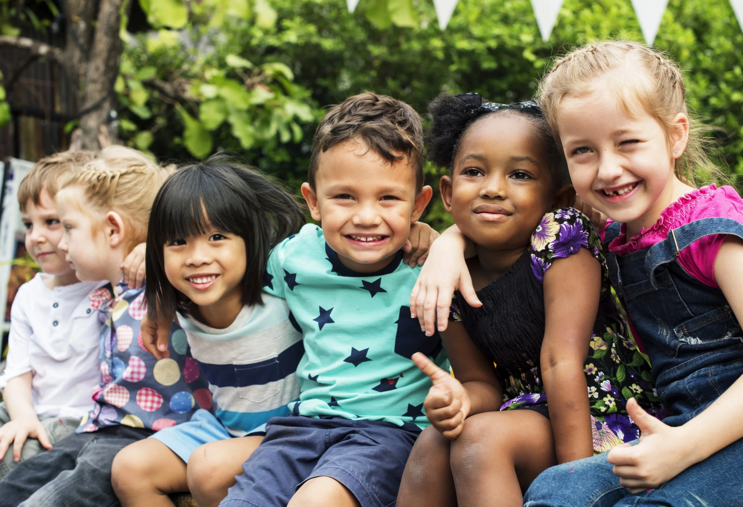 A diverse group of children sitting close together outdoors with greenery in the background, smiling and posing for the photo.