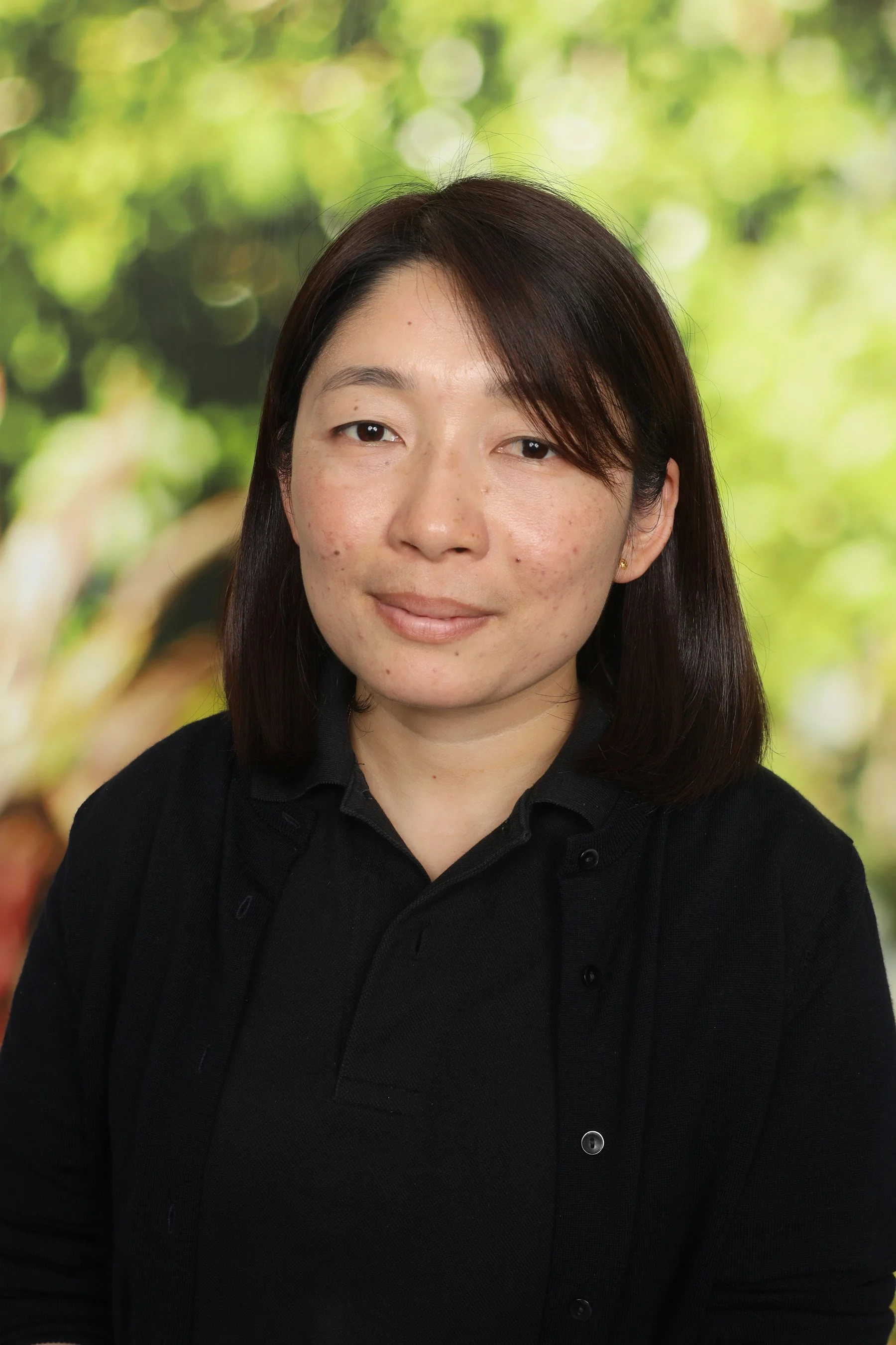 Portrait of a woman with dark brown hair, wearing a black shirt, against a blurred green outdoor background.