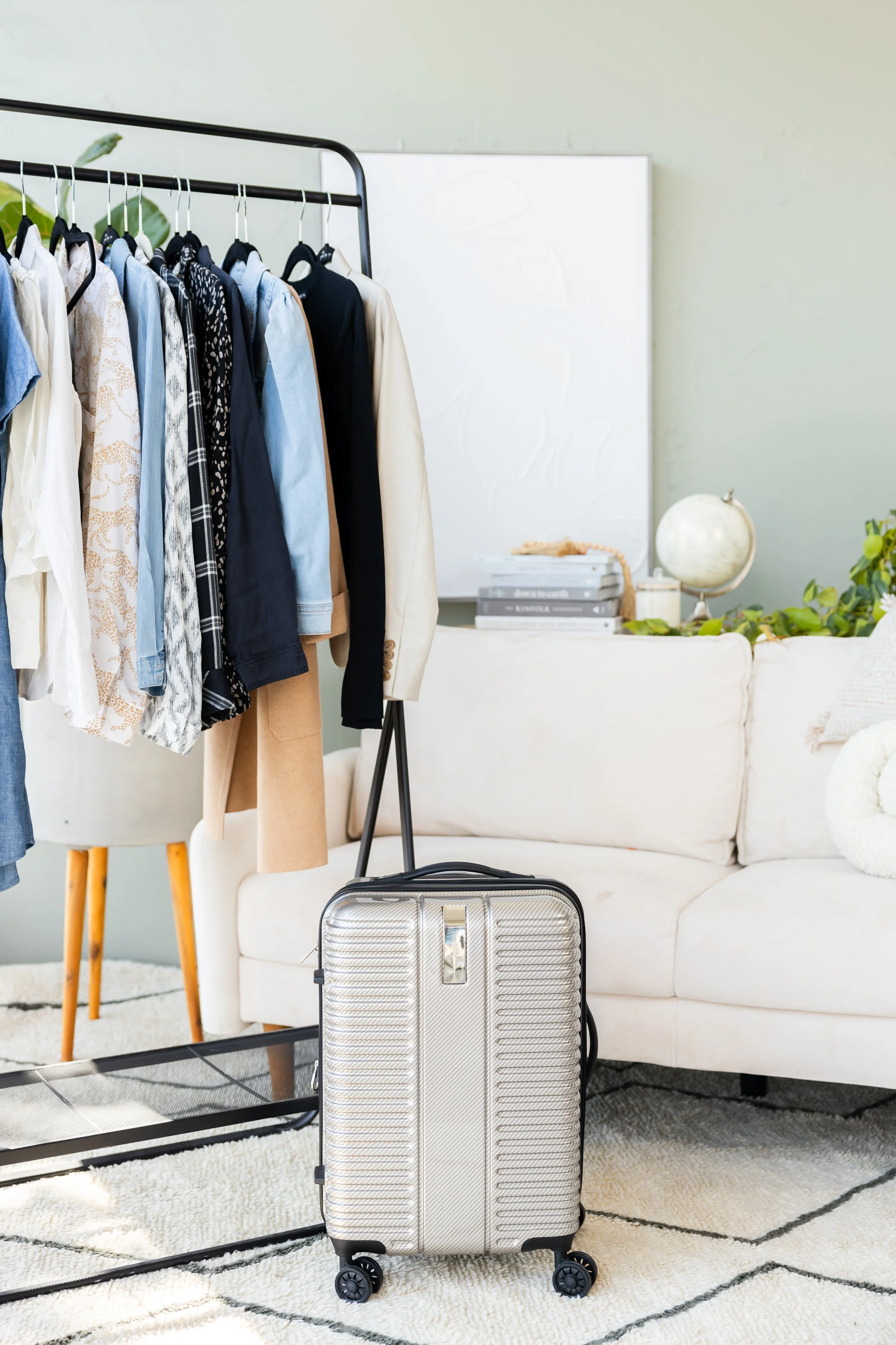 A beige suitcase with black wheels in front of a clothing rack filled with various shirts and jackets, set in a living room with a white sofa, a globe, and a small stack of books.