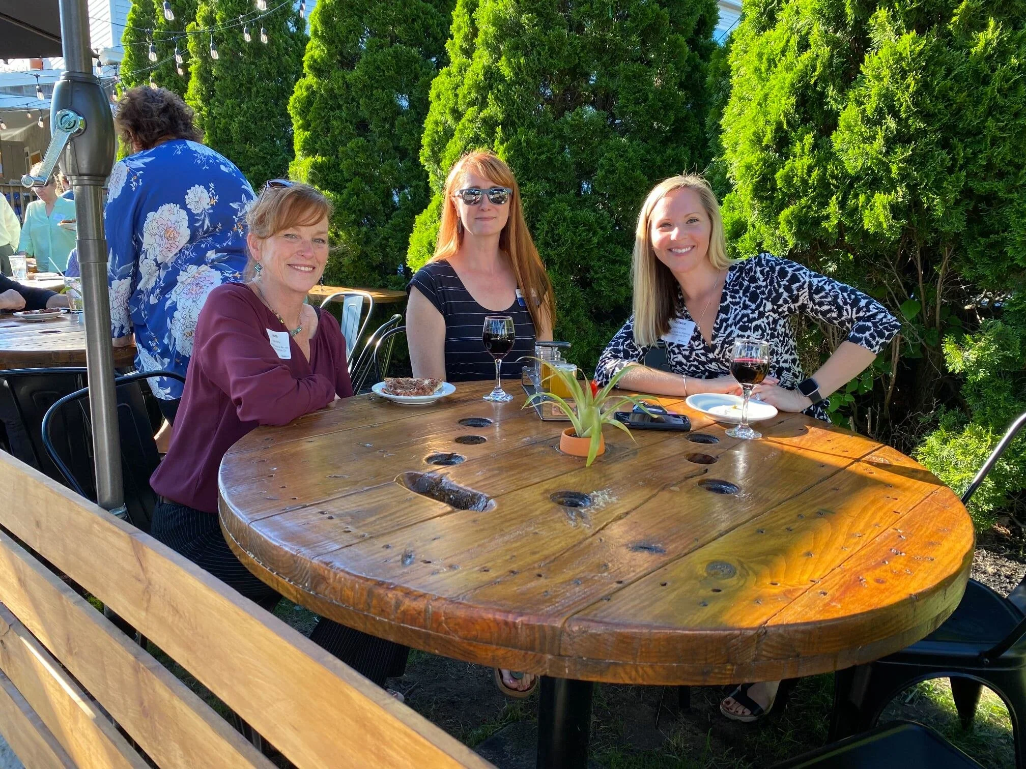 Three women sitting at a wooden outdoor table with food and drinks, smiling at the camera, with greenery in the background.