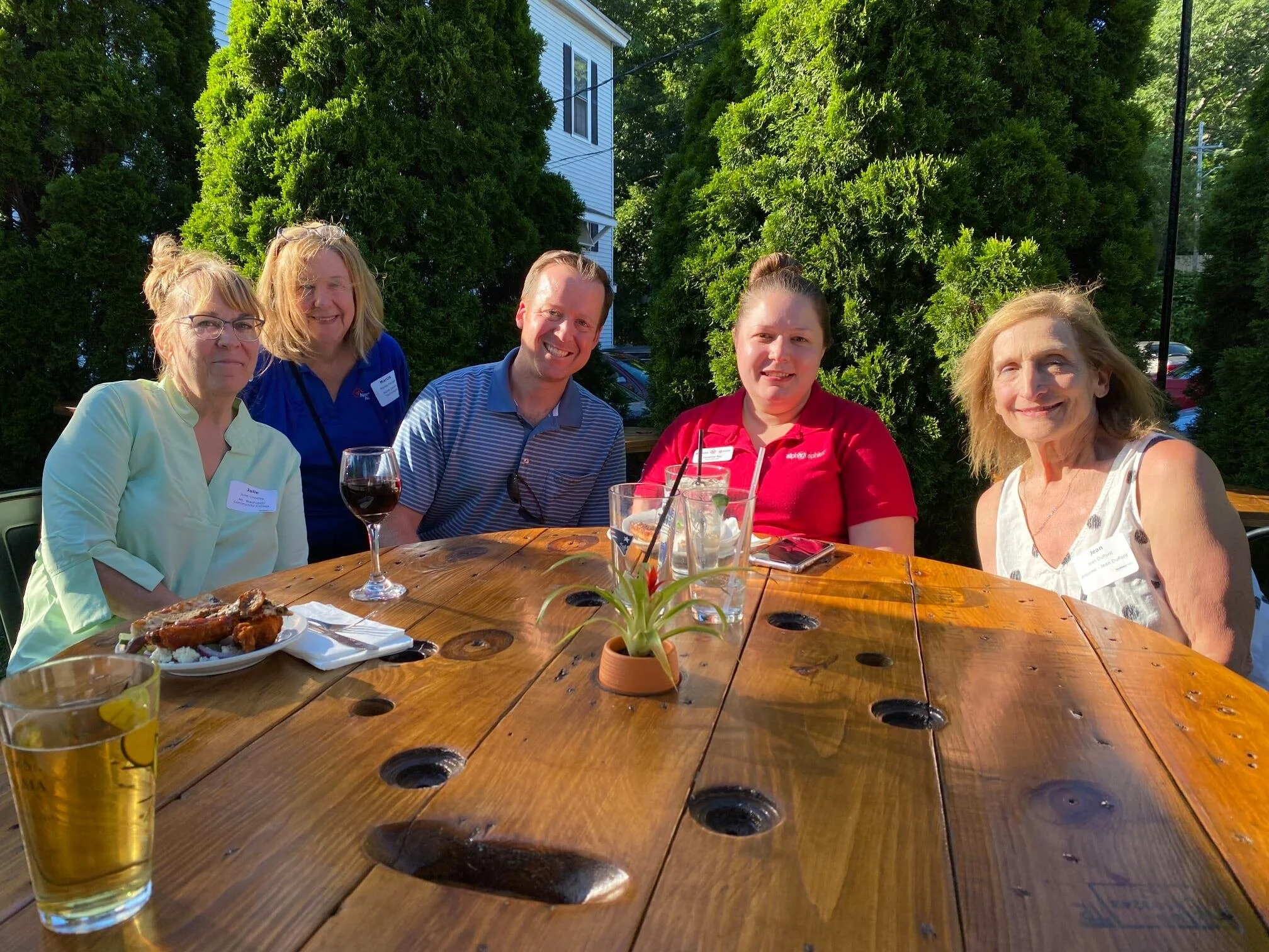 Group of five people sitting at an outdoor wooden table with drinks and food, smiling, surrounded by greenery in a backyard setting.