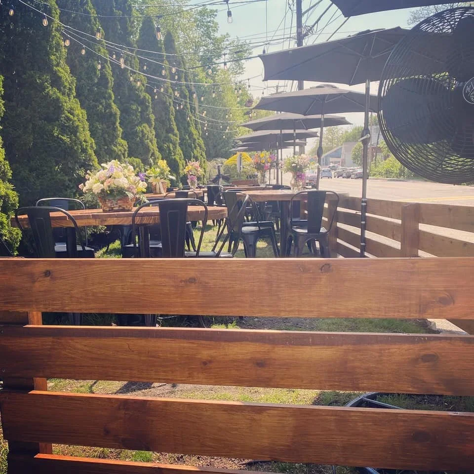 Outdoor dining area with wooden tables, black chairs, large black umbrellas, and flower arrangements in vases, adjacent to a wooden fence and greenery, under string lights.