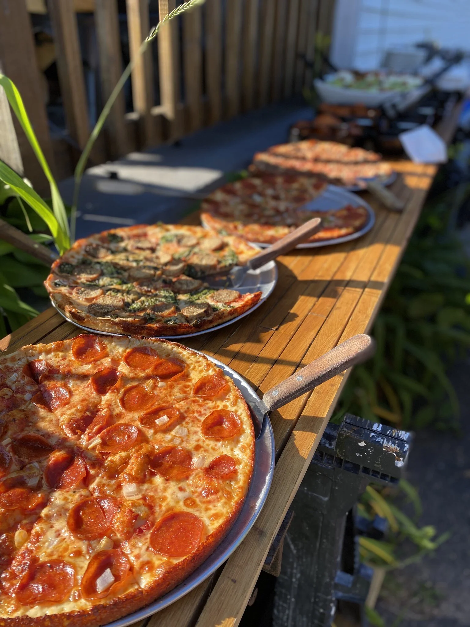 Several pizza pies with toppings like pepperoni and mushrooms on a wooden table outdoors with a fence and plants in the background.
