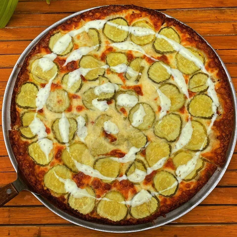 A pizza with pickles and drizzled with white sauce on a wooden surface.