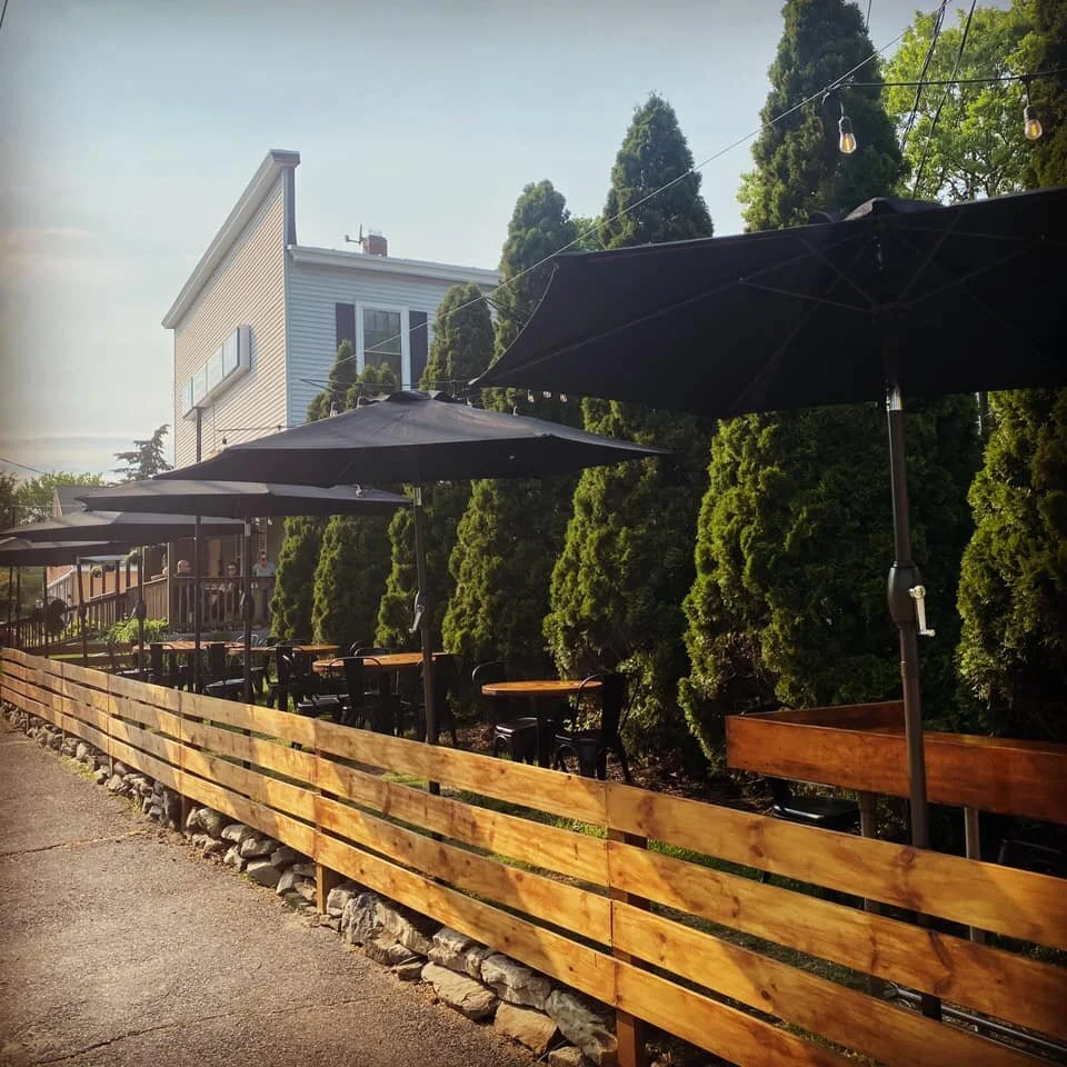 Outdoor patio with black umbrellas, wooden tables, black chairs, wooden fence, lush green bushes, and string lights, with a house in the background.