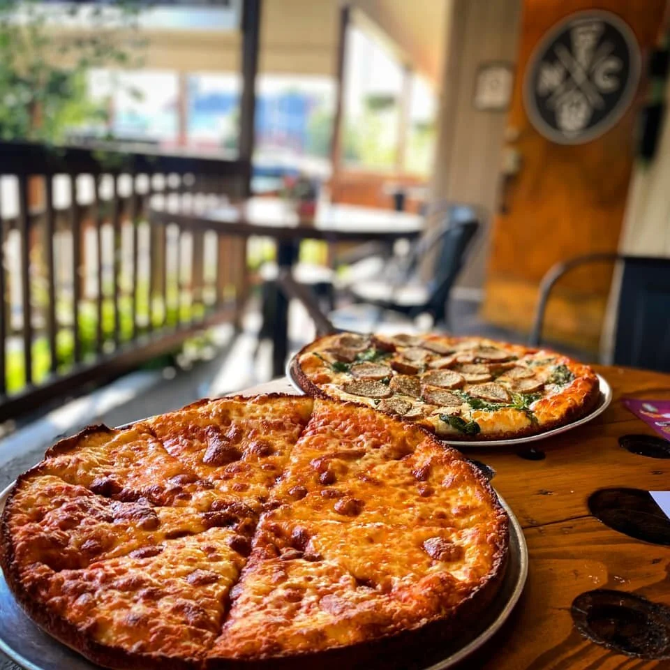 Two pizzas on a wooden table inside a restaurant, with a balcony and outdoor seating visible in the background.