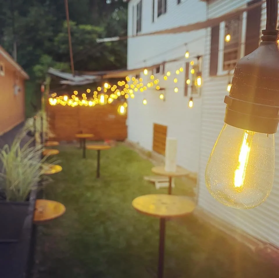 Outdoor patio with string lights, wooden picnic tables, a small wooden shed, and string lights hanging against a white house exterior during dusk.