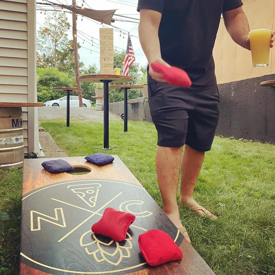 Person standing outdoors playing cornhole, holding a glass of orange juice, with a cornhole board and bean bags in front of him.