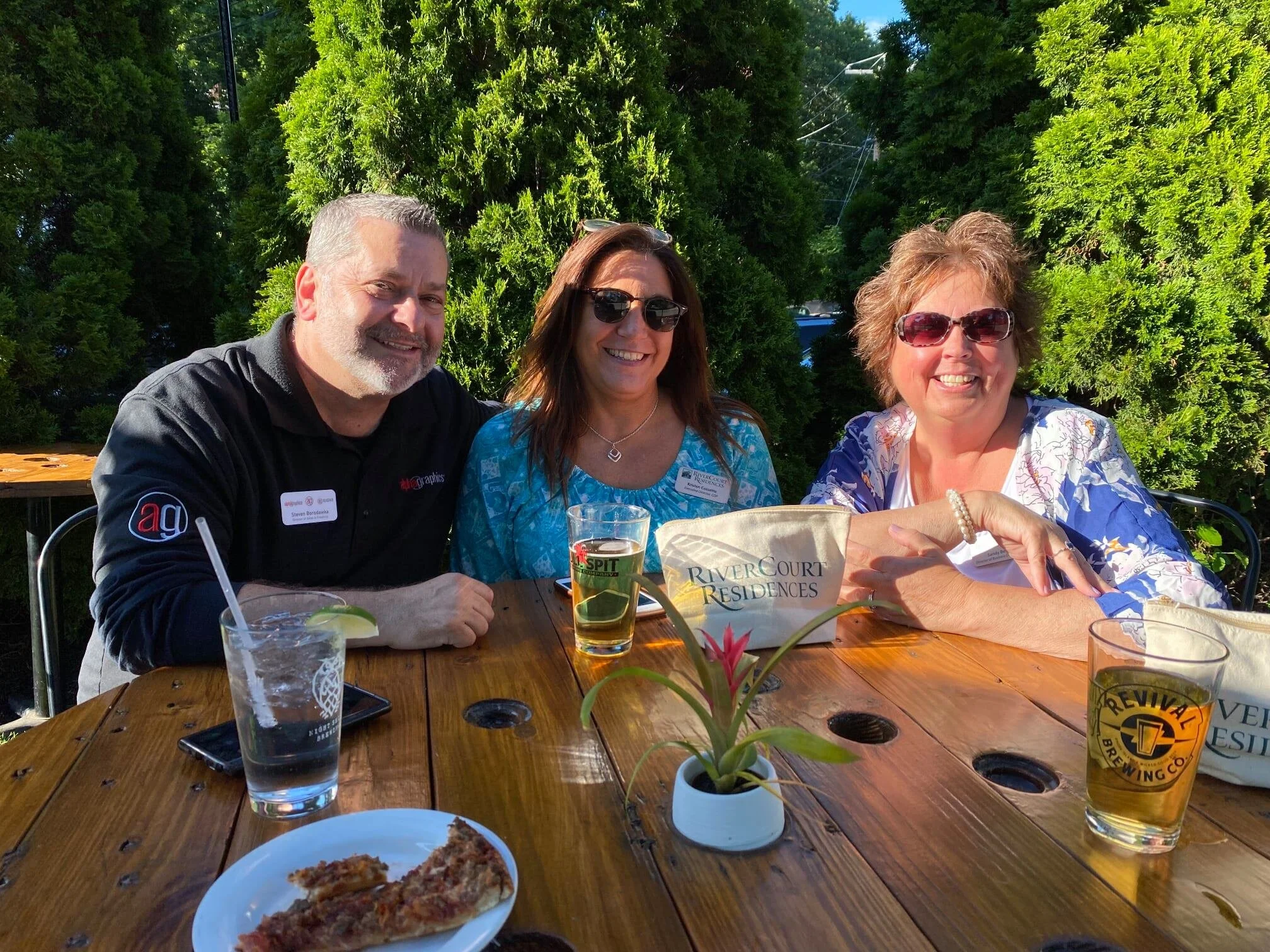 Three people sitting at a wooden table outside, smiling. There are drinks, food, and a small potted plant on the table, with green trees in the background.