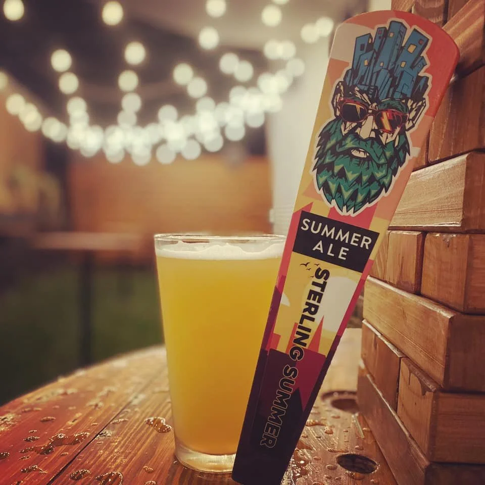 A glass of yellow summer ale beer next to a colorful label featuring a bearded man with sunglasses, set on a wooden table with string lights in the background.