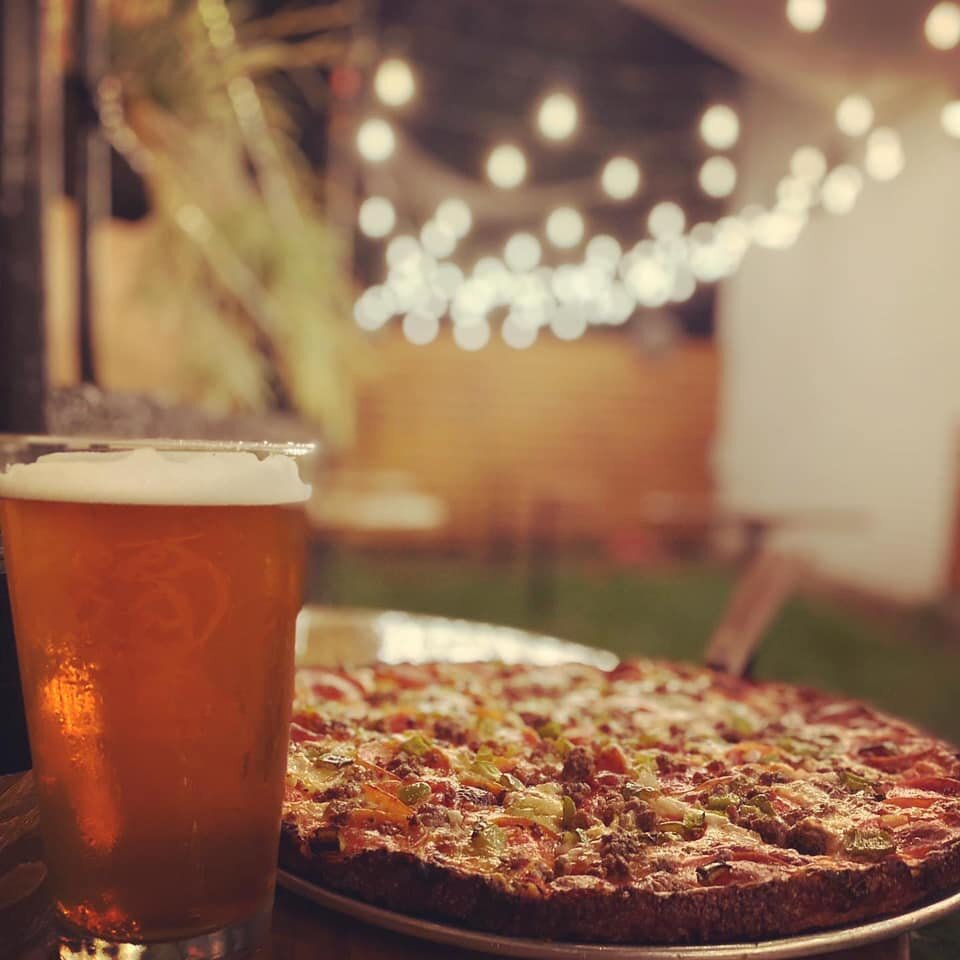 A pizza with toppings including sausage, pepperoni, and vegetables next to a glass of beer on a table in an outdoor setting with string lights in the background.