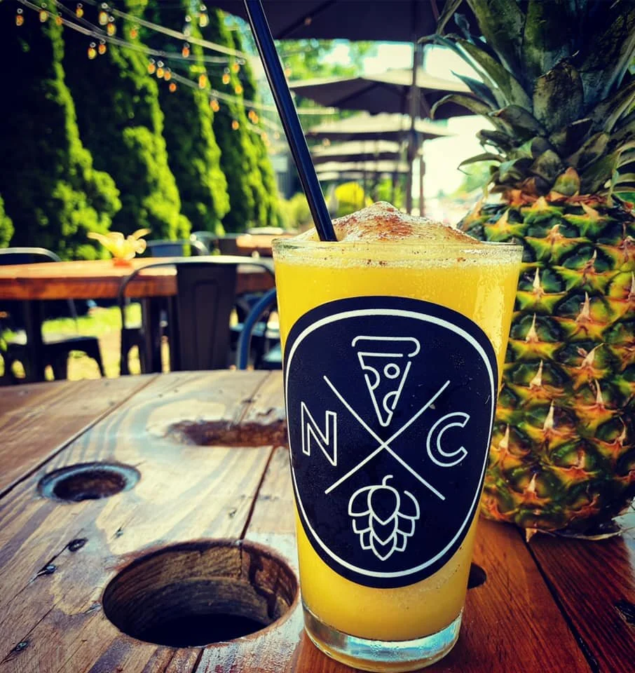 A tall glass of yellow frozen drink with a straw on a wooden table outdoors, with a pineapple and green plants in the background.