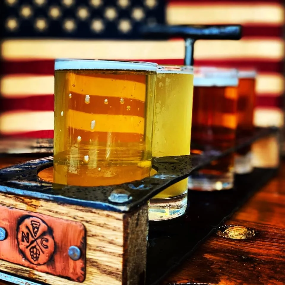 Three glasses of beer on a wooden flight tray, with an American flag in the background.