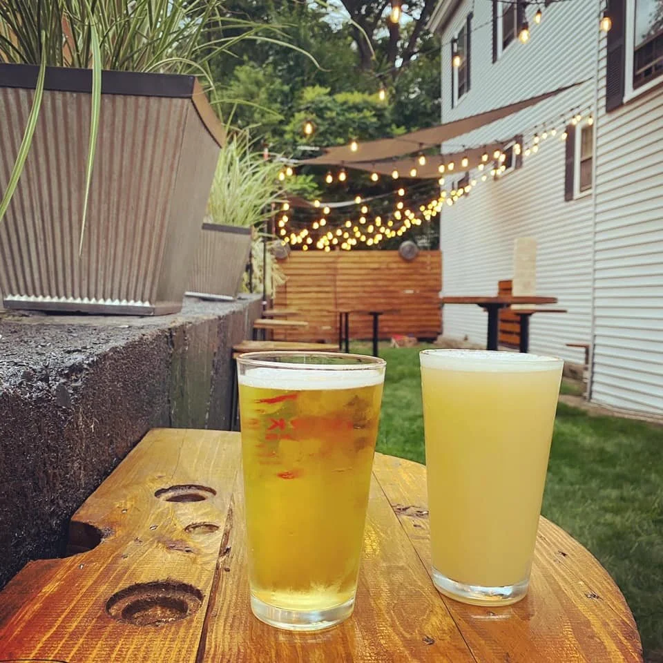 Two glasses of pale-colored beverages placed on a wooden outdoor table, with string lights and a white house in the background.