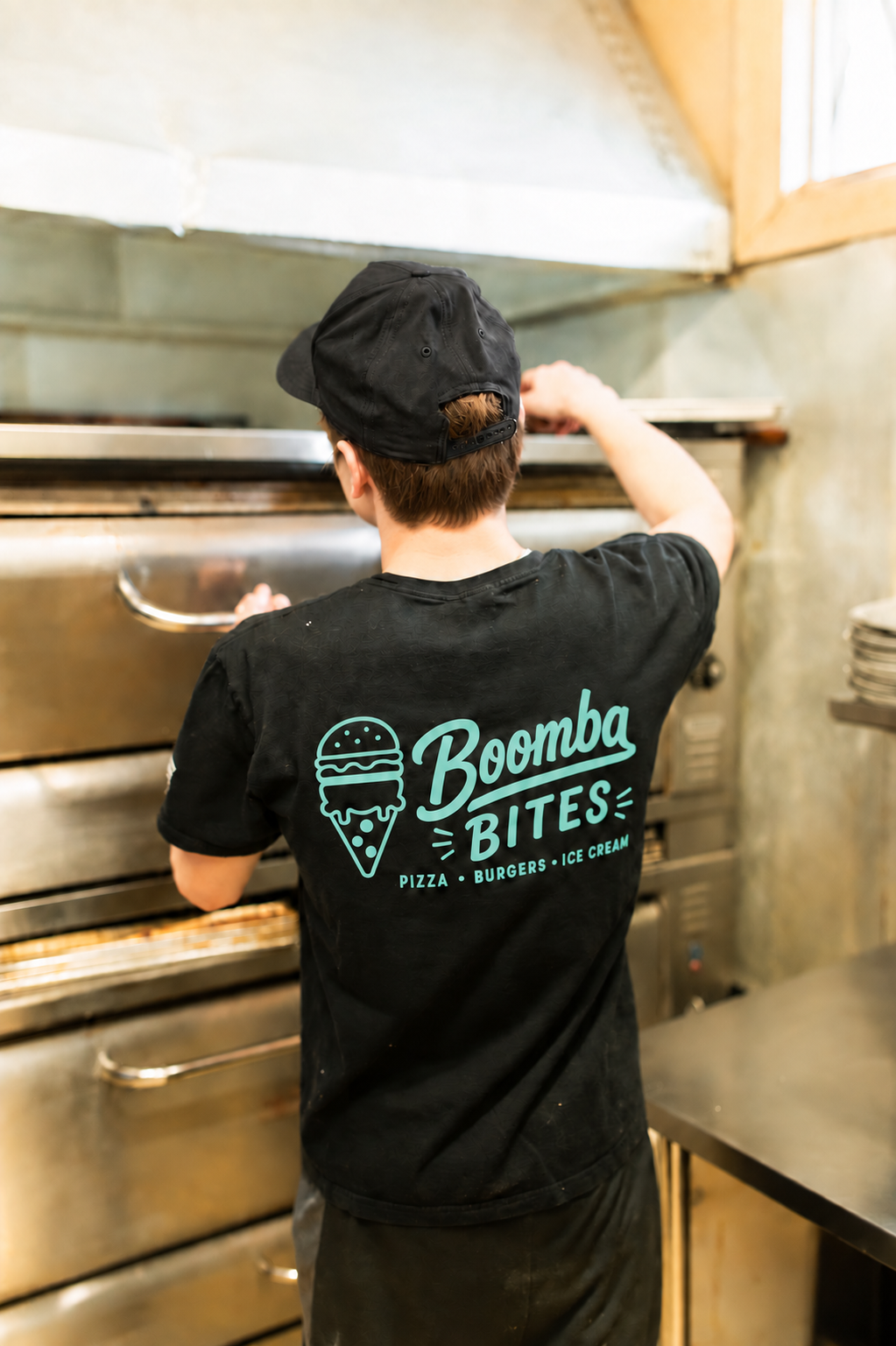 A person with short brown hair wearing a black cap and black T-shirt, with the logo "Boomba Bites" on the back, is cooking in a commercial kitchen using a large oven.