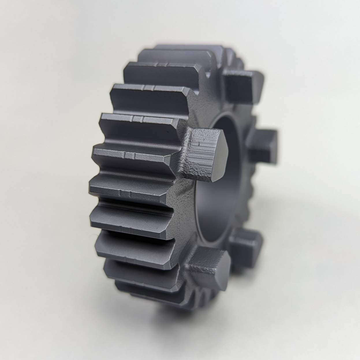 A metal gear with evenly spaced teeth, seen from a side angle on a plain white surface, coated in molybdenum disulfide.