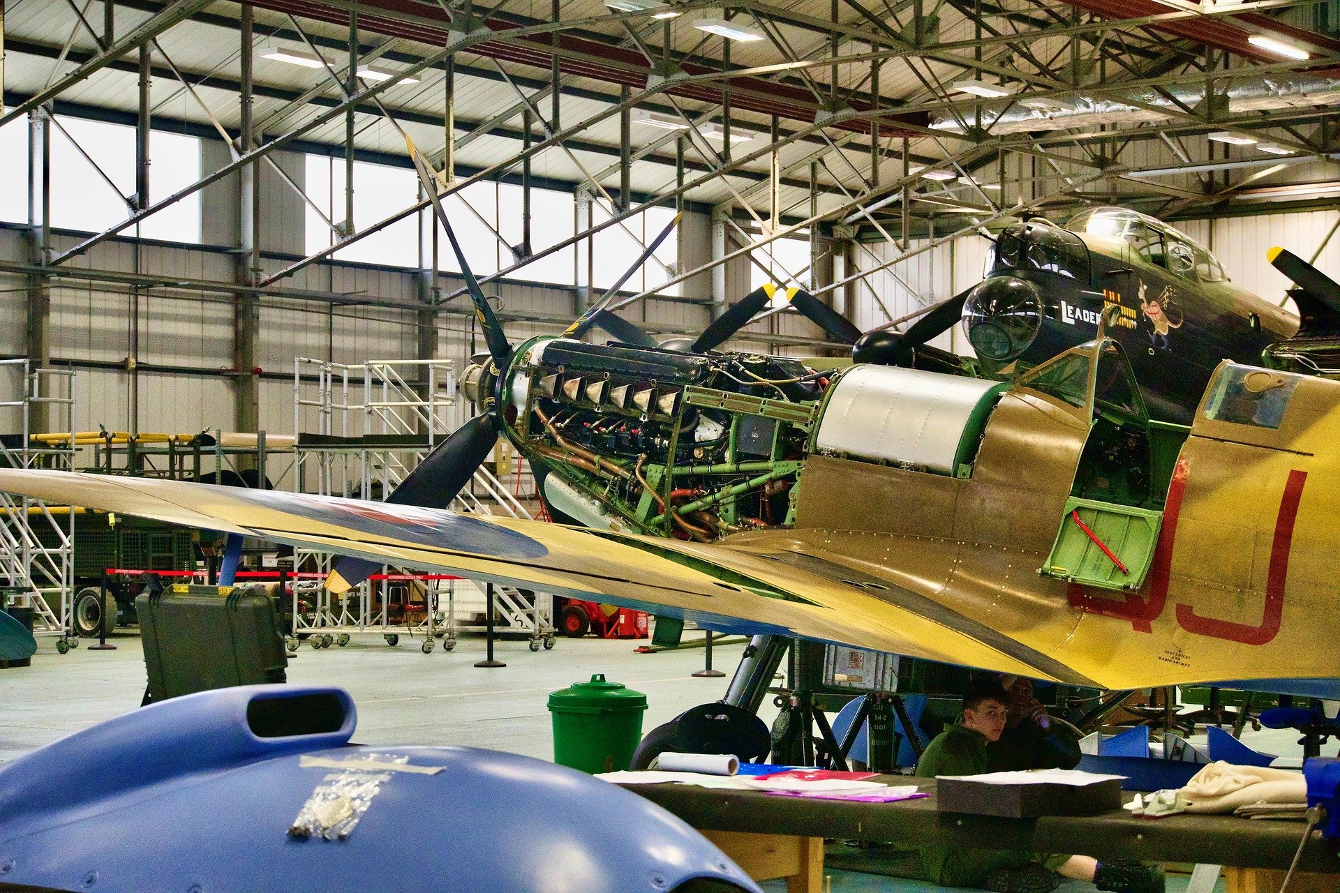 Inside an aircraft hangar with vintage aircraft, including a partially disassembled plane with an exposed engine, a black aircraft with artwork and markings, and a yellow plane. Various equipment, scaffolding, and a person sitting at a table are visible.