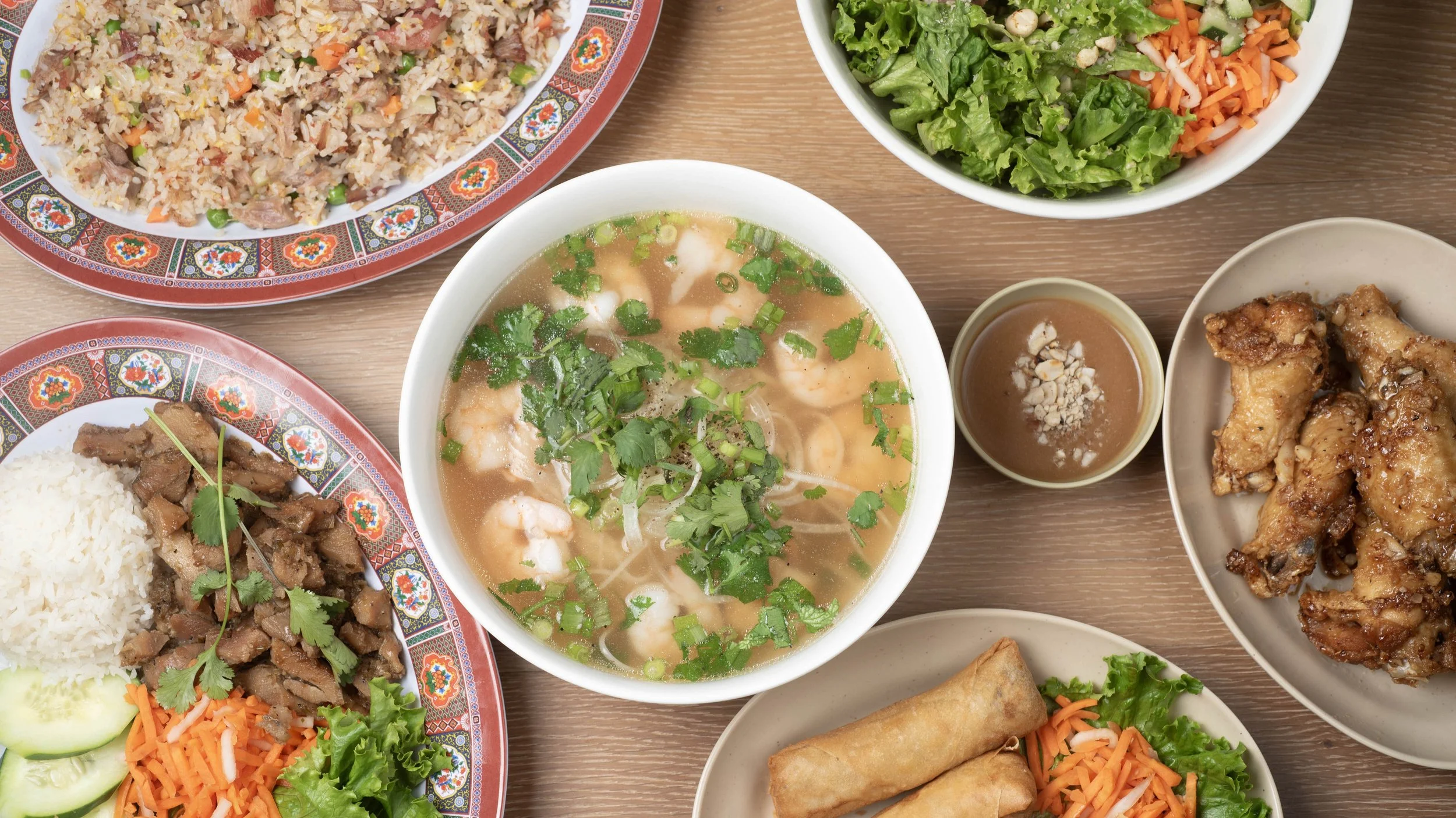 Assorted dishes including fried rice, salad, shrimp soup, spring rolls, fried chicken, and beef served on decorative plates and bowls on a wooden table.