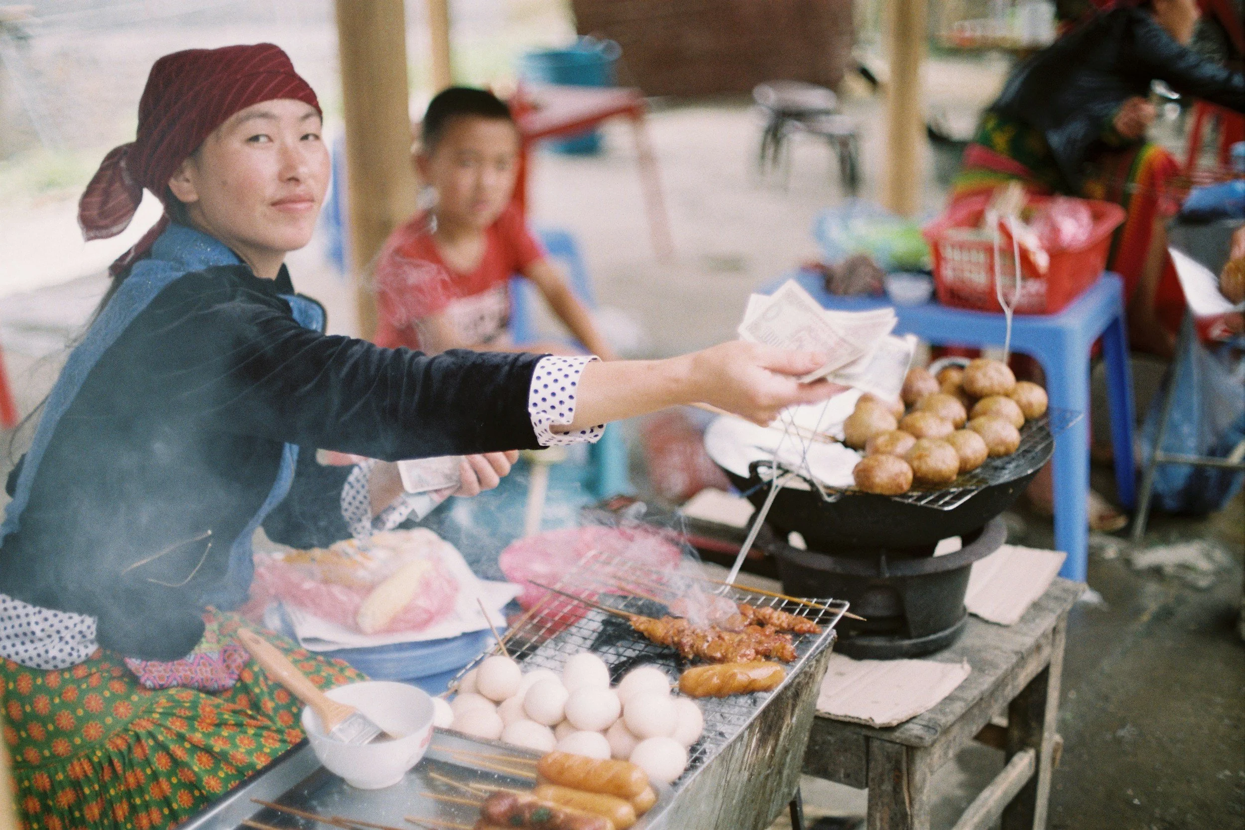 A woman selling grilled meat, eggs, and sausages at a street food stall, with a young girl and other people in the background.