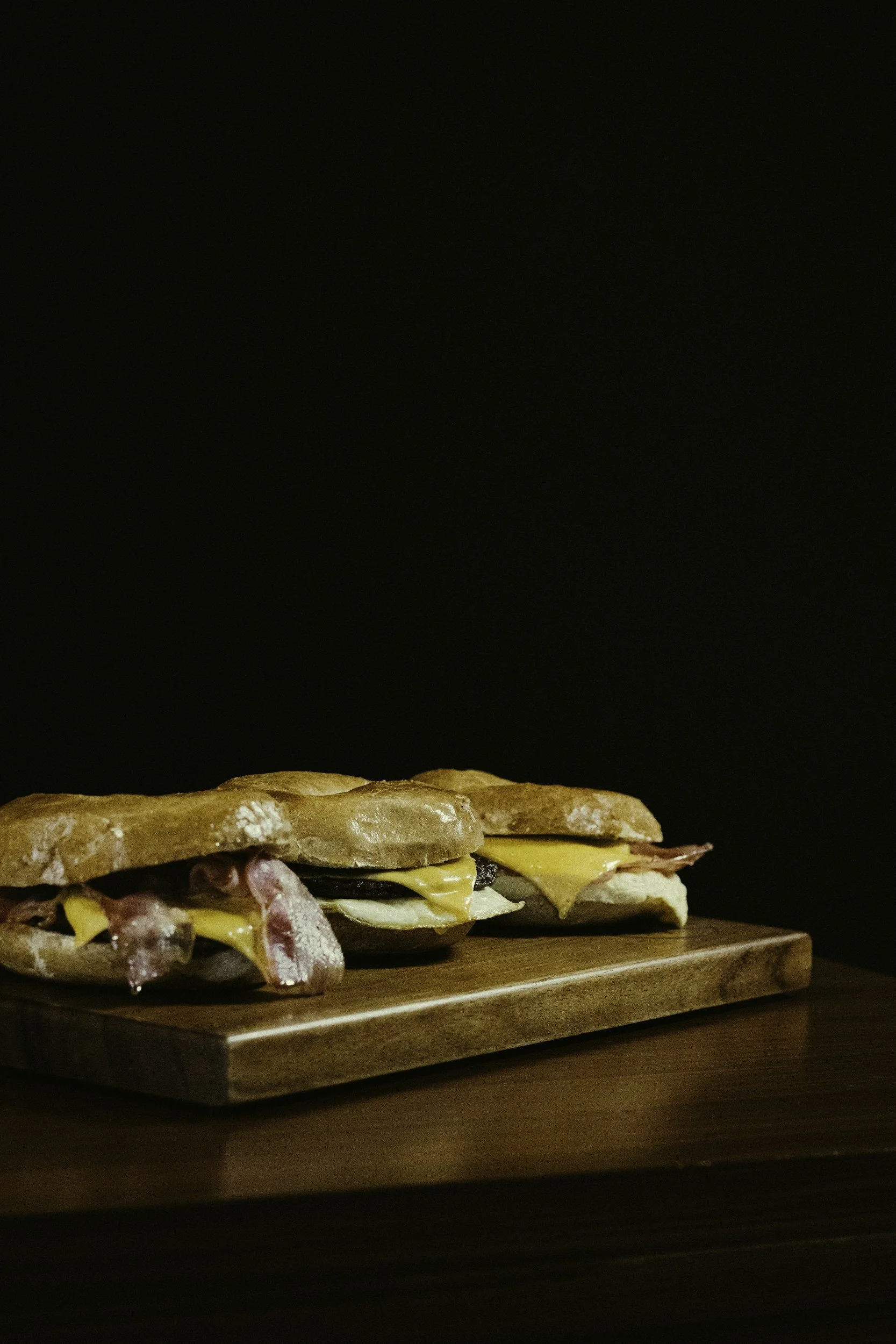 Three breakfast sandwiches with bacon, cheese, egg, and meats on toasted bread, placed on a wooden cutting board against a dark background.