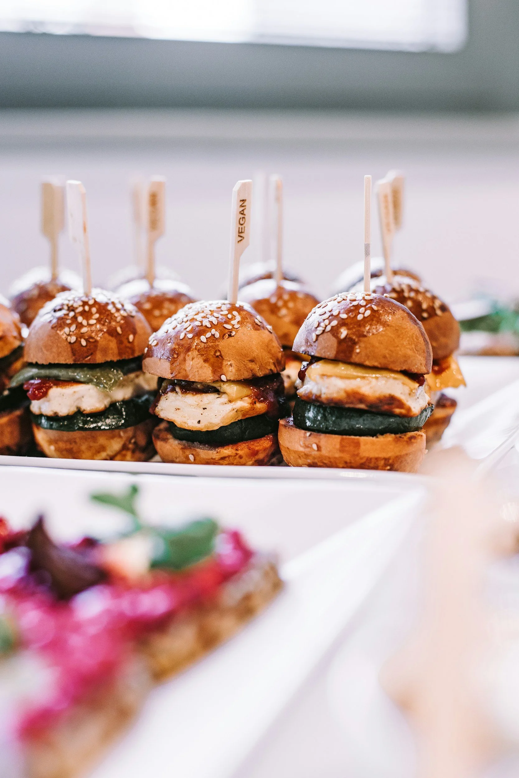 Mini vegetarian sliders with sesame seed buns, filled with grilled vegetables and cheese, topped with toothpicks labeled 'VEGAN'.
