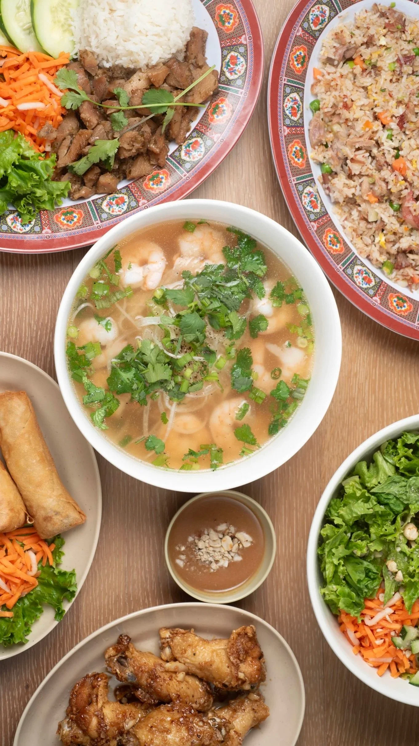 A variety of Asian dishes including a bowl of shrimp soup garnished with cilantro, plates of fried rice and stir-fried beef with vegetables, spring rolls, and a salad with lettuce and shredded carrots, all arranged on a wooden table.