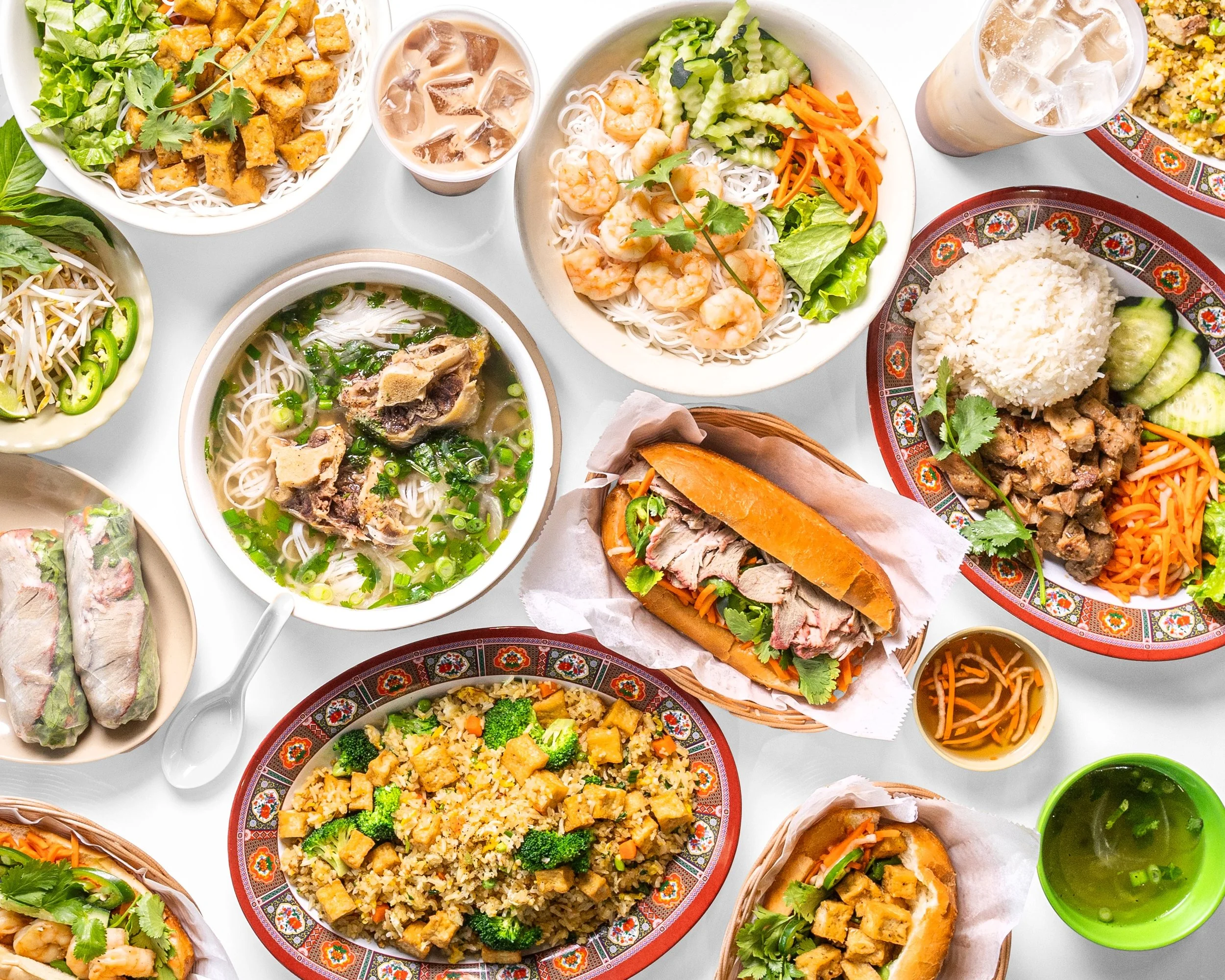 A variety of Asian dishes on a white table, including noodle soups, spring rolls, fried rice, banh mi sandwiches, shrimp salad, and iced drinks.