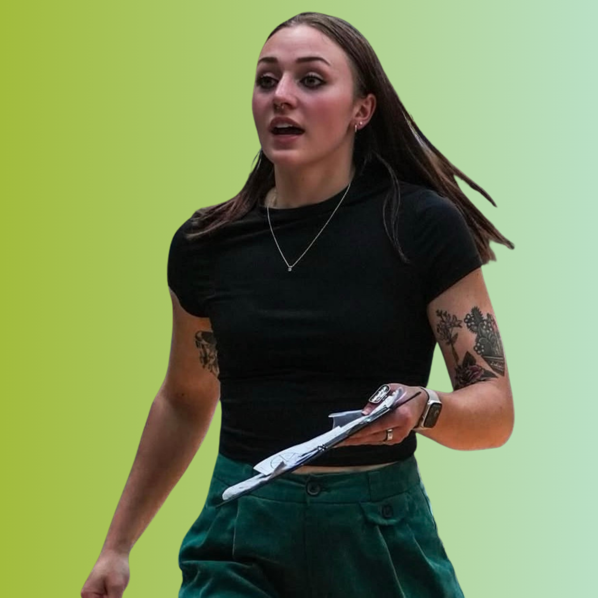 A young woman with long brown hair and tattoos on both arms, wearing a black t-shirt, green pants, a necklace, a smartwatch, and holding a phone and papers, walking against a green gradient background.