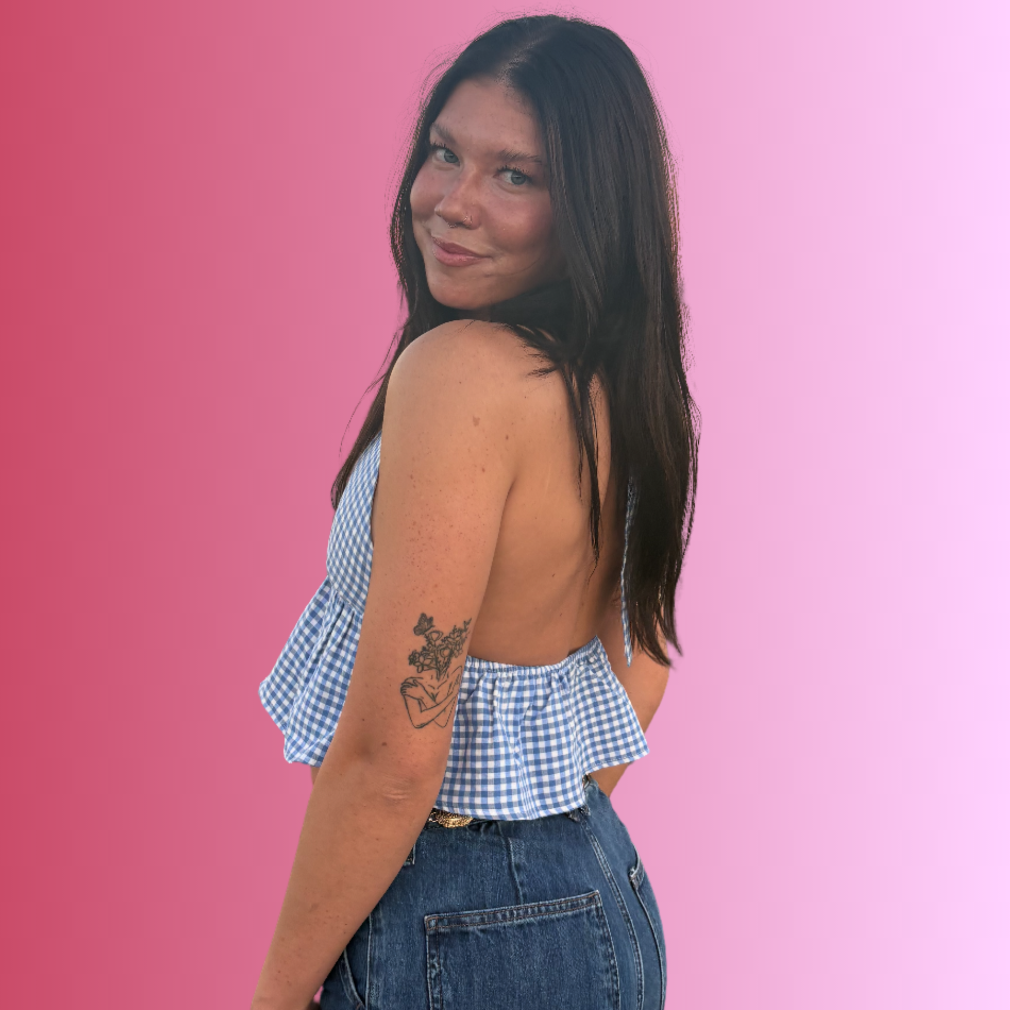 A woman with long dark hair and a tattoo on her left arm standing against a pink and purple gradient background, looking over her shoulder with a slight smile, wearing a blue and white checkered sleeveless top and jeans.
