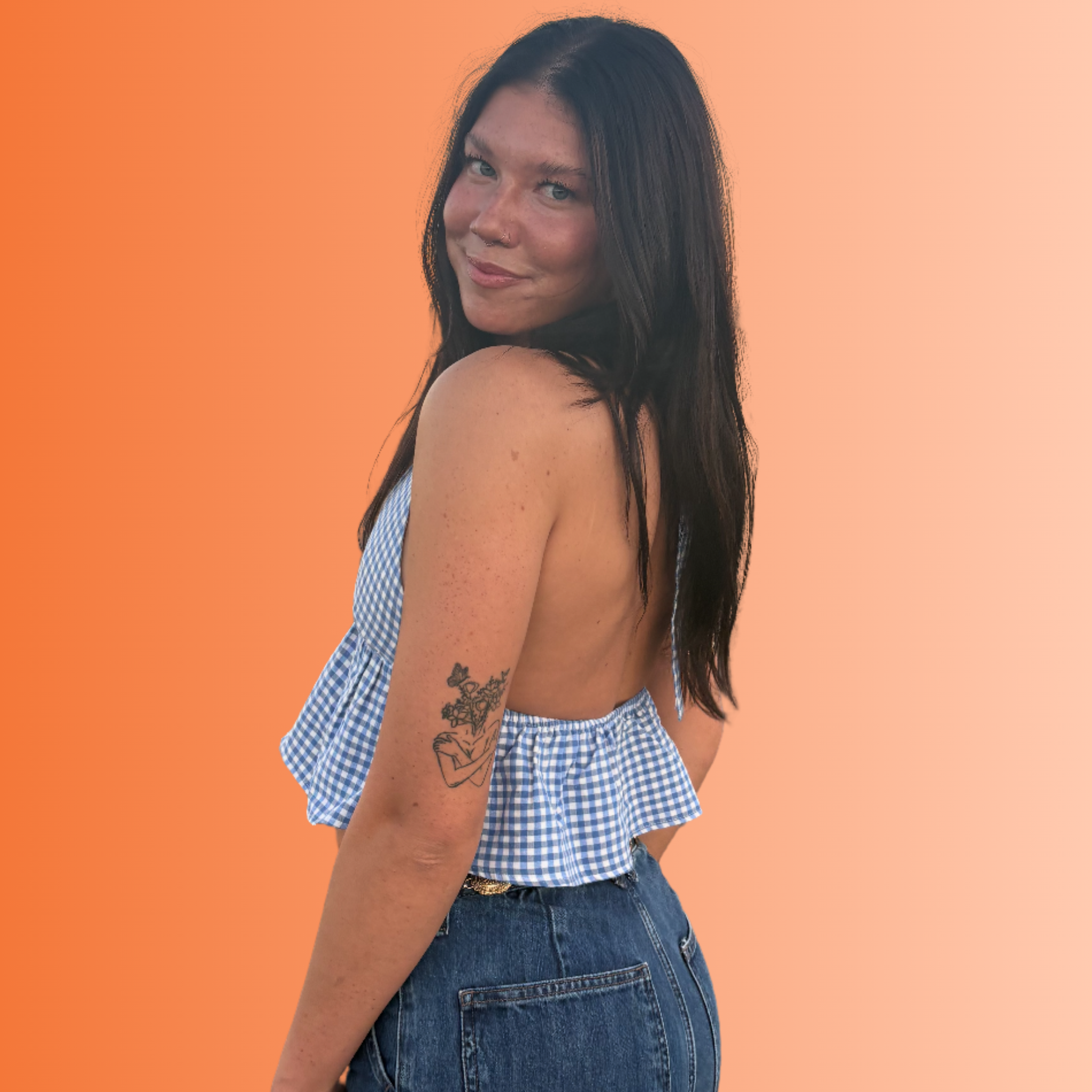 A woman with dark hair and freckles, wearing a backless blue gingham top and jeans, standing against a gradient orange background.