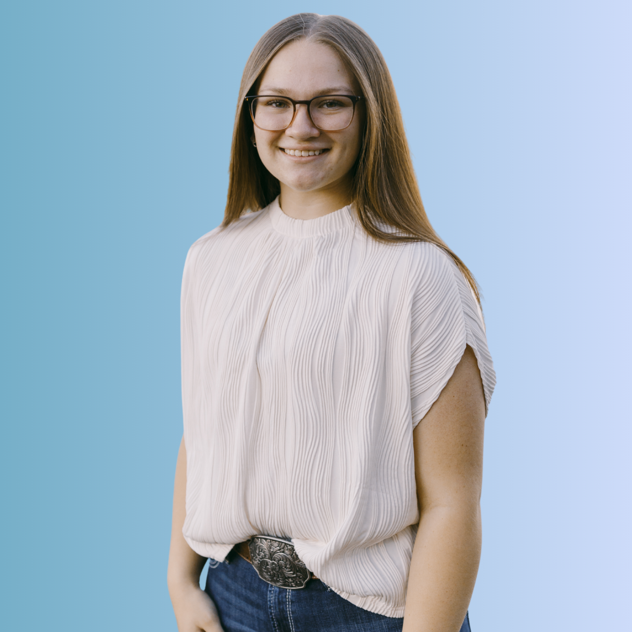 A young woman with long brown hair, wearing glasses, a white pleated blouse, and jeans, smiling against a blue background.