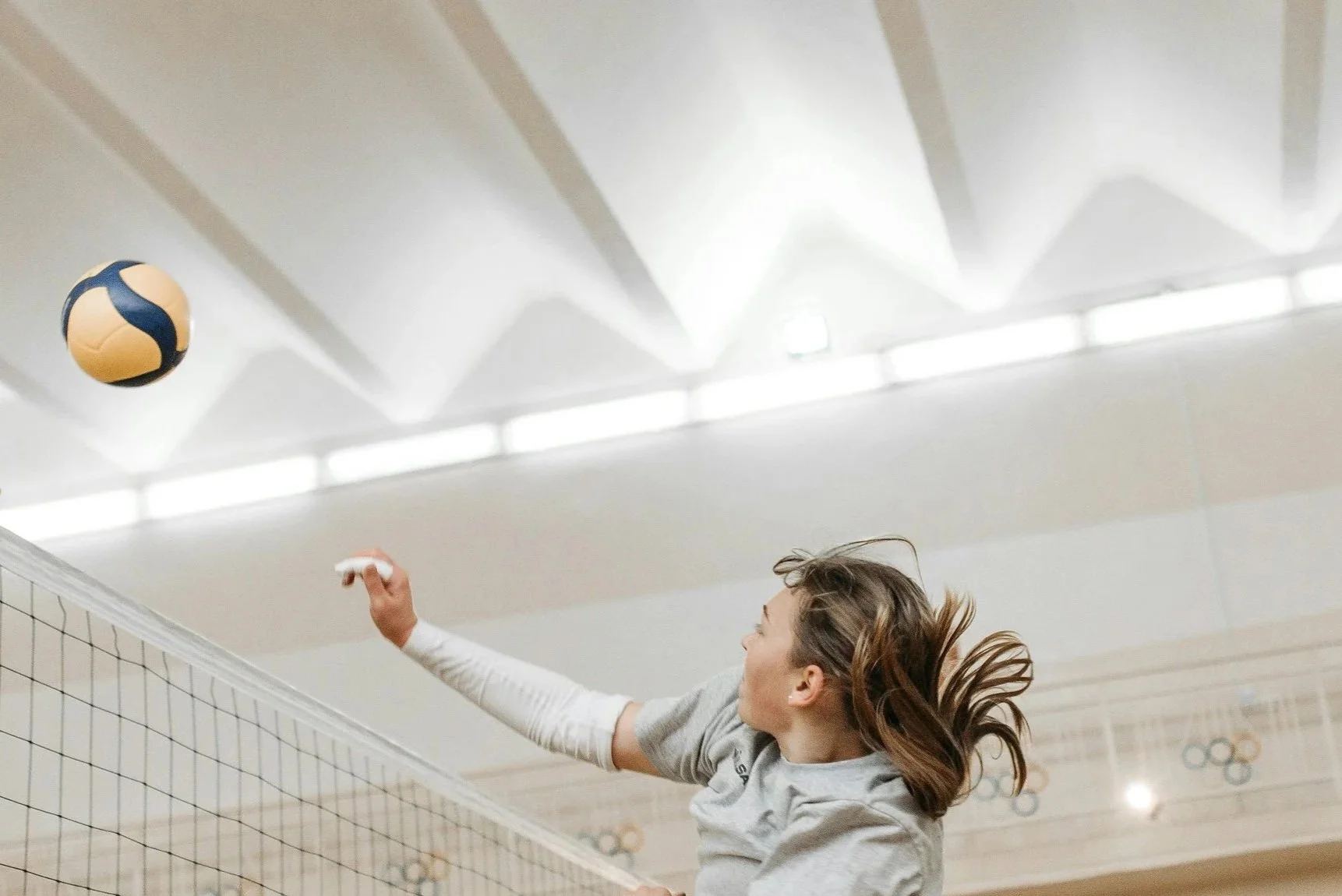 A young woman playing volleyball indoors, reaching to hit a volleyball near the net, with her hair flowing.